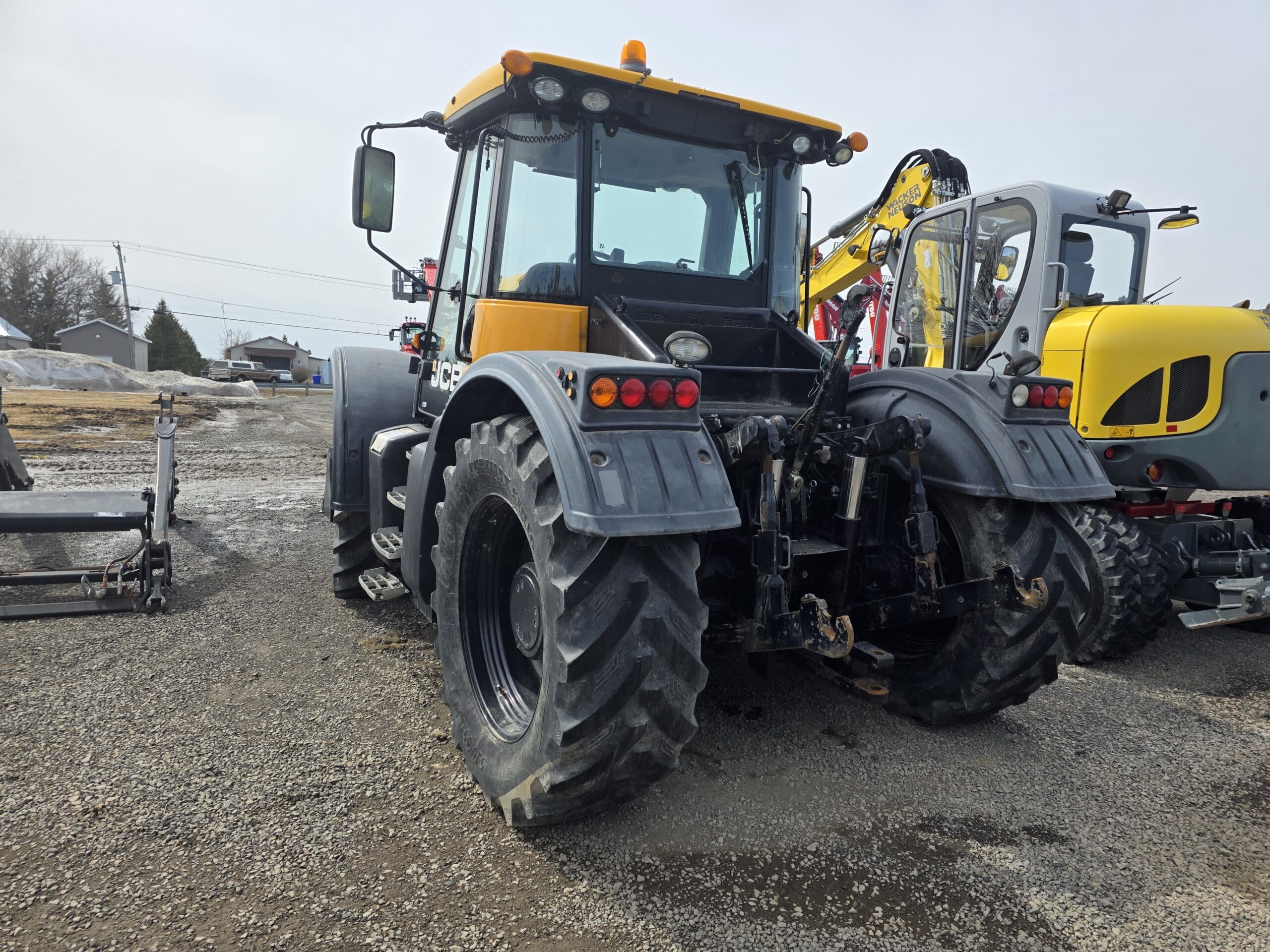 Un tracteur jaune et noir est garé sur un terrain en gravier, vu de l'arrière. Un autre engin lourd est visible à droite. Le ciel est couvert et quelques bâtiments apparaissent au loin.