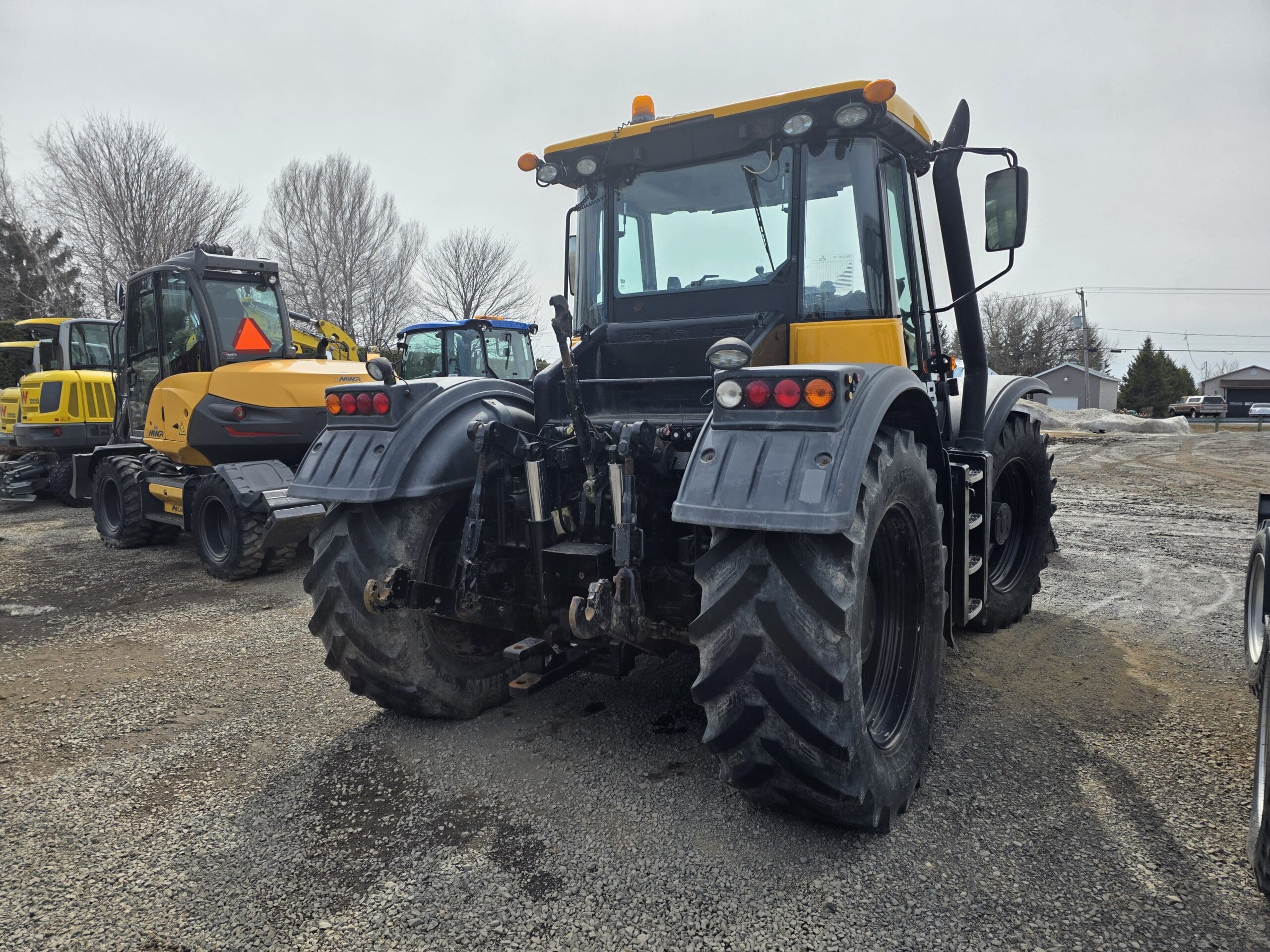 Un gros tracteur noir et jaune est garé sur un terrain en gravier, avec d'autres engins de chantier à proximité, par une journée nuageuse. Des arbres et quelques bâtiments sont visibles en arrière-plan.