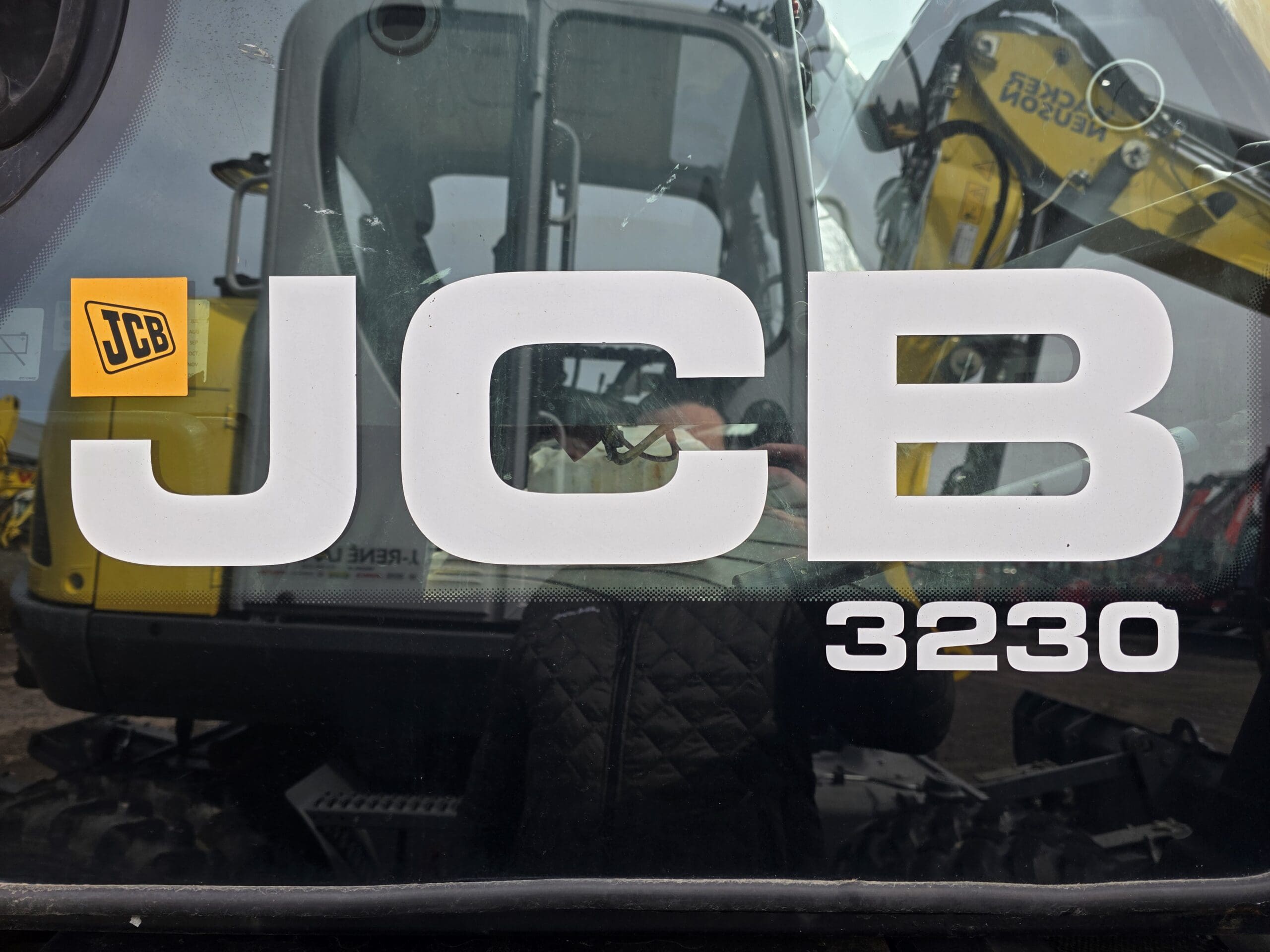 Gros plan du côté d'une machinerie lourde affichant de grandes lettres blanches « JCB 3230 » avec un logo JCB jaune ; des reflets et des parties d'autres équipements de construction sont visibles en arrière-plan.