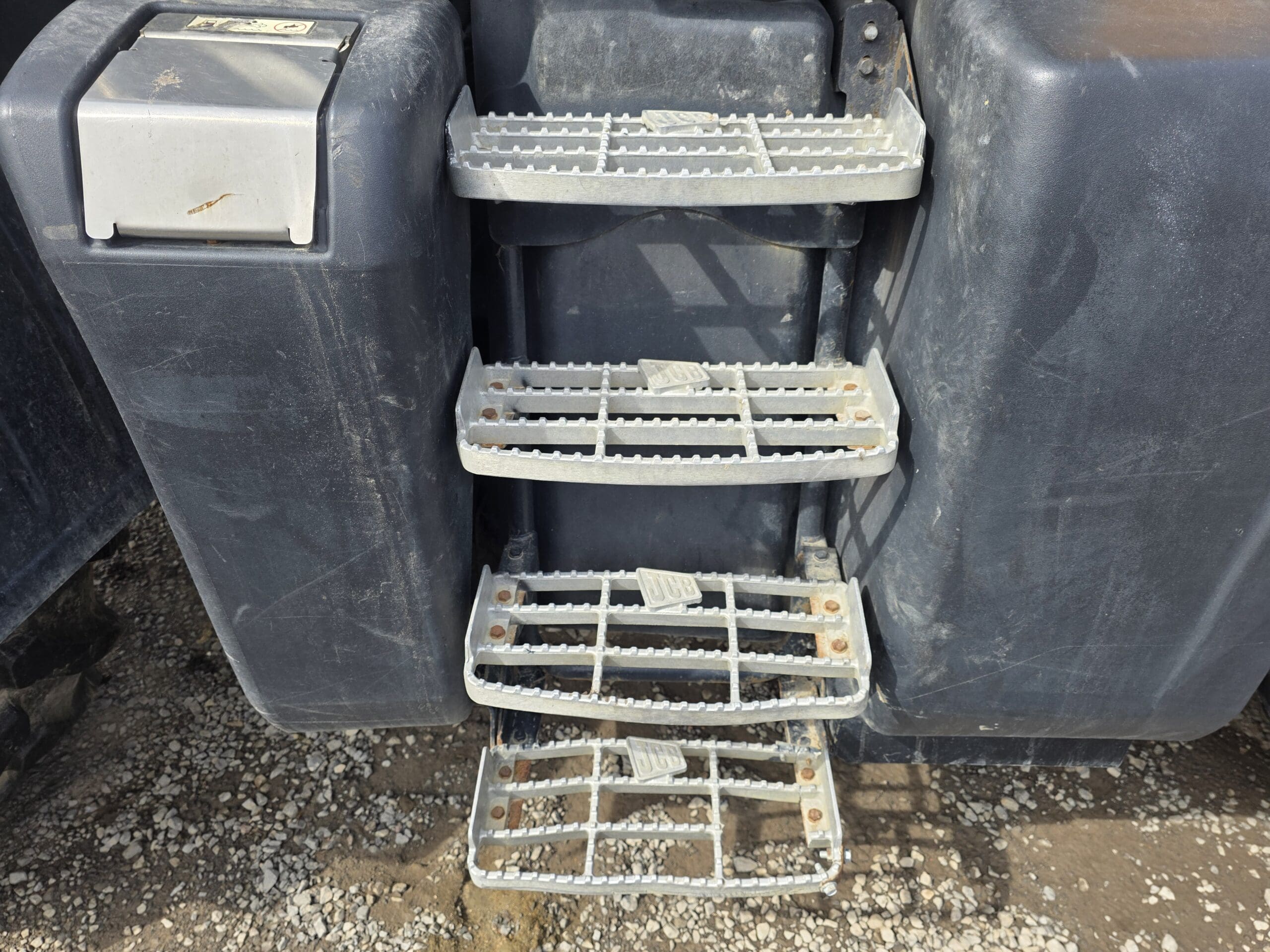 Quatre marchepieds métalliques fixés à un grand véhicule industriel sombre, avec de la rouille visible sur les boulons. Les marchepieds sont placés au-dessus du gravier et de la terre. Le côté gauche comporte un compartiment ou couvercle gris.