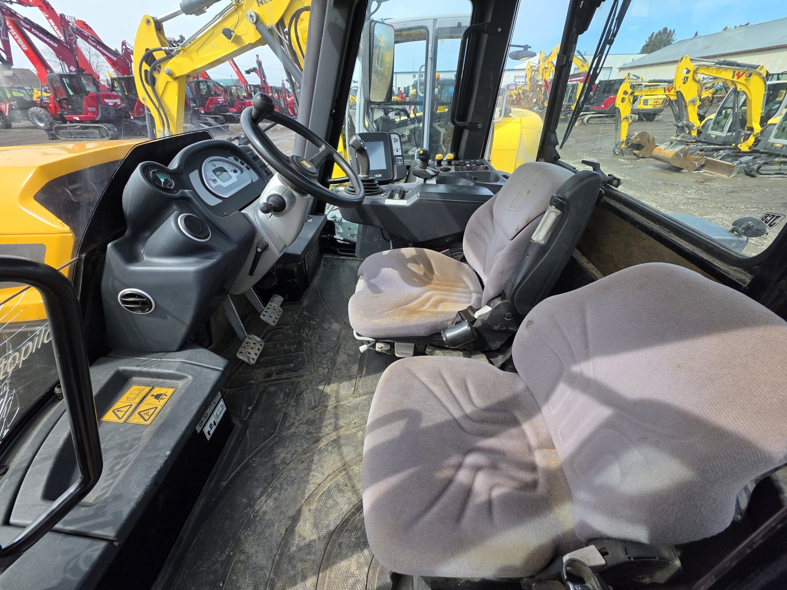 Vue intérieure d'une cabine de véhicule de construction montrant deux sièges en tissu, un volant, un panneau de commande et divers leviers, avec plusieurs excavatrices jaunes visibles à l'extérieur à travers les fenêtres.