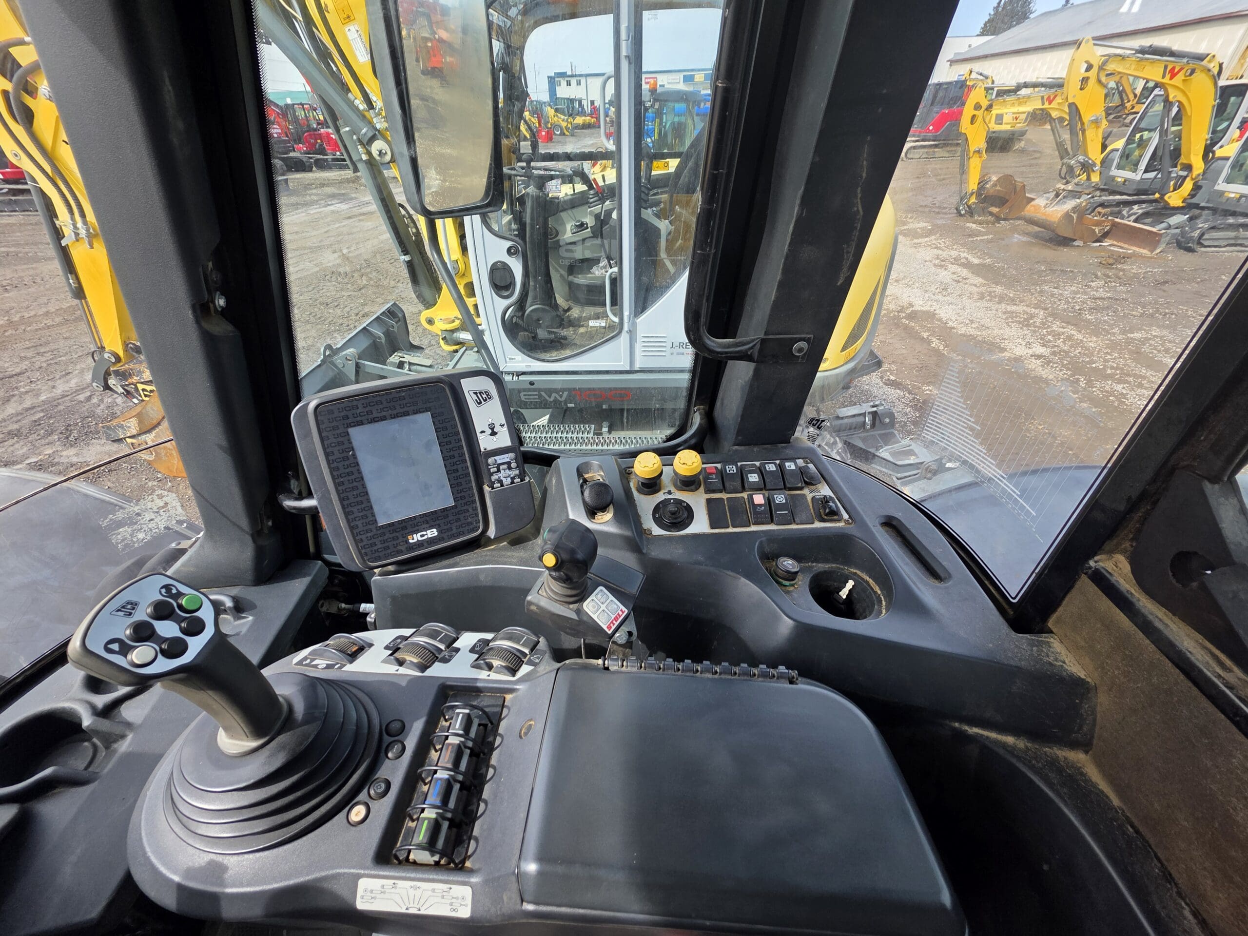 Vue de l'intérieur d'une cabine d'excavatrice, montrant le panneau de commande, le joystick, les boutons et l'affichage numérique, avec des engins de construction visibles à l'extérieur à travers de grandes fenêtres.
