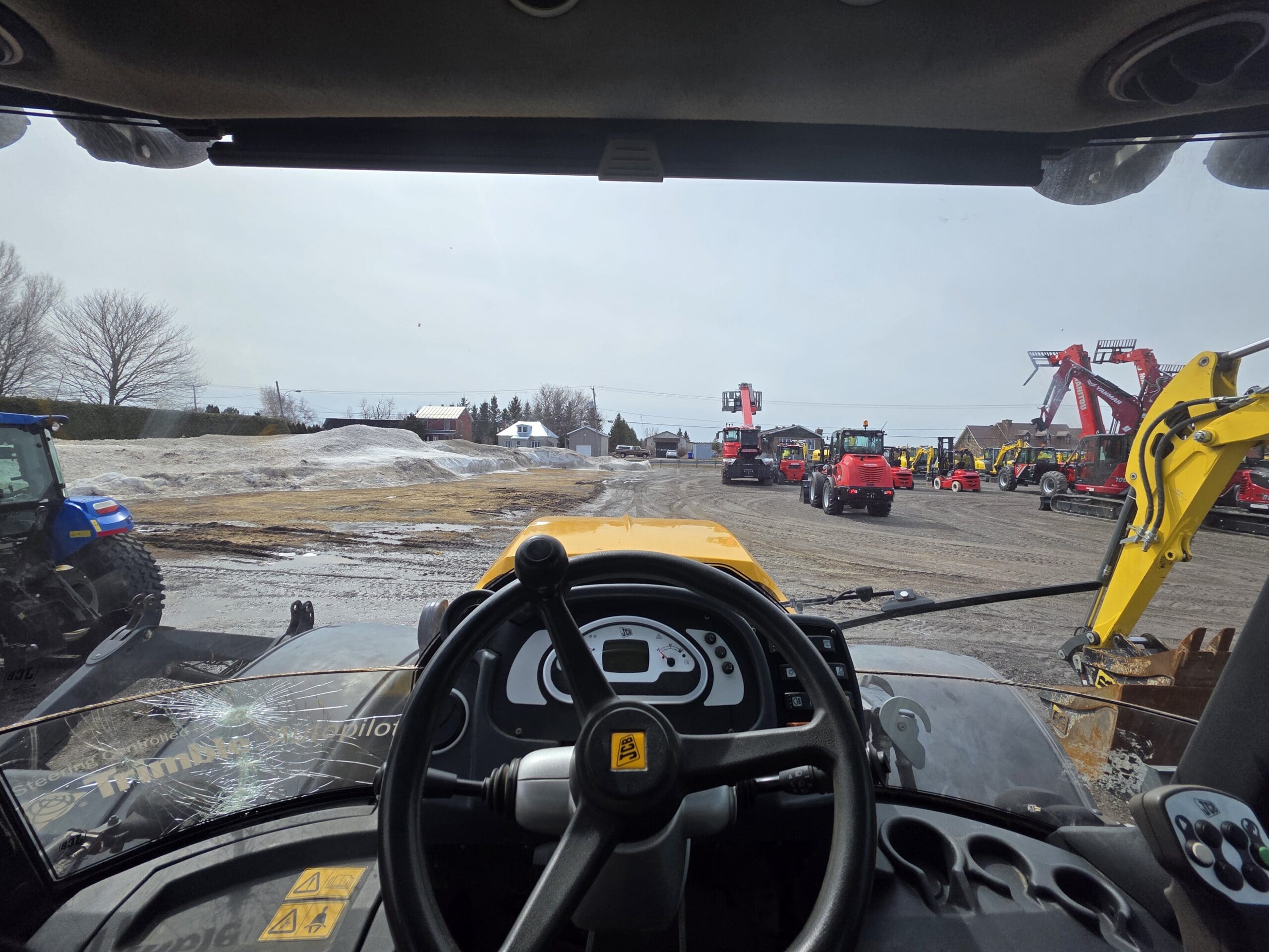 Vue de l'intérieur de la cabine d'un tracteur, montrant le volant et le tableau de bord. À l'extérieur, divers engins de chantier et agricoles sont garés sur un terrain en gravier, avec des plaques de neige et quelques arbres en arrière-plan.