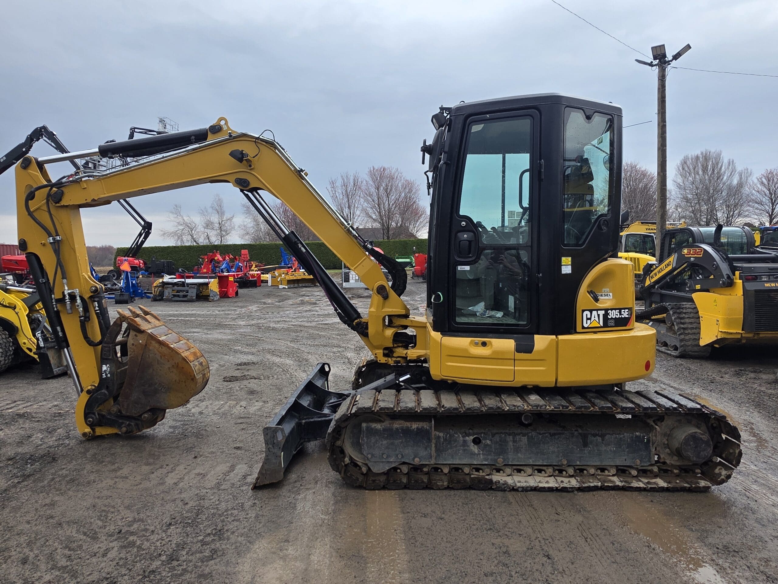 Une mini-pelle jaune Caterpillar 305.5E avec un godet de creusement est garée sur un terrain en gravier, entourée d'autres équipements de construction sous un ciel nuageux.