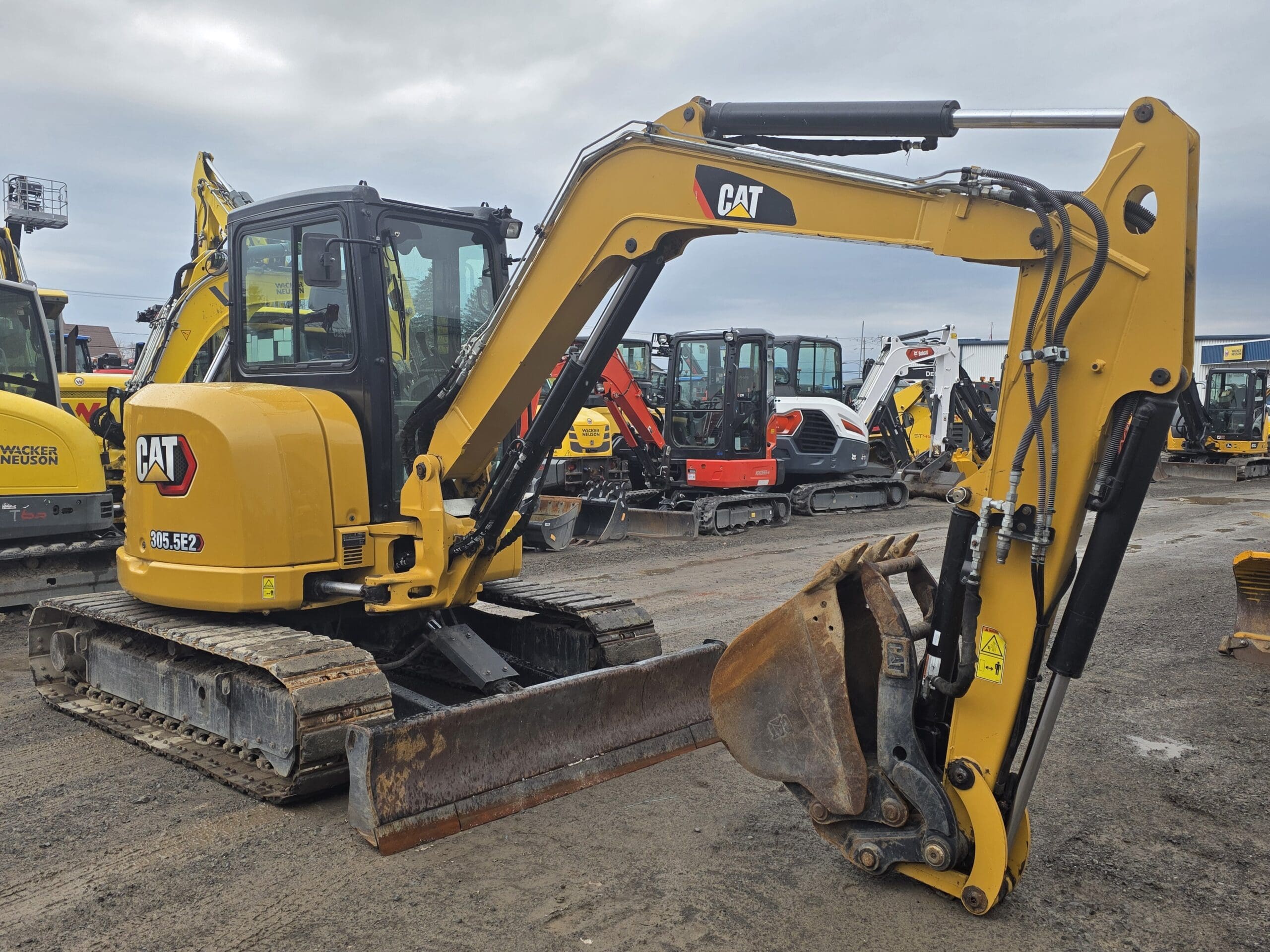 Une mini-pelle CAT 303.5E2 jaune est garée sur un terrain vague parmi d'autres engins de chantier, sous un ciel nuageux. Le bras et le godet de la pelle sont orientés vers le sol.