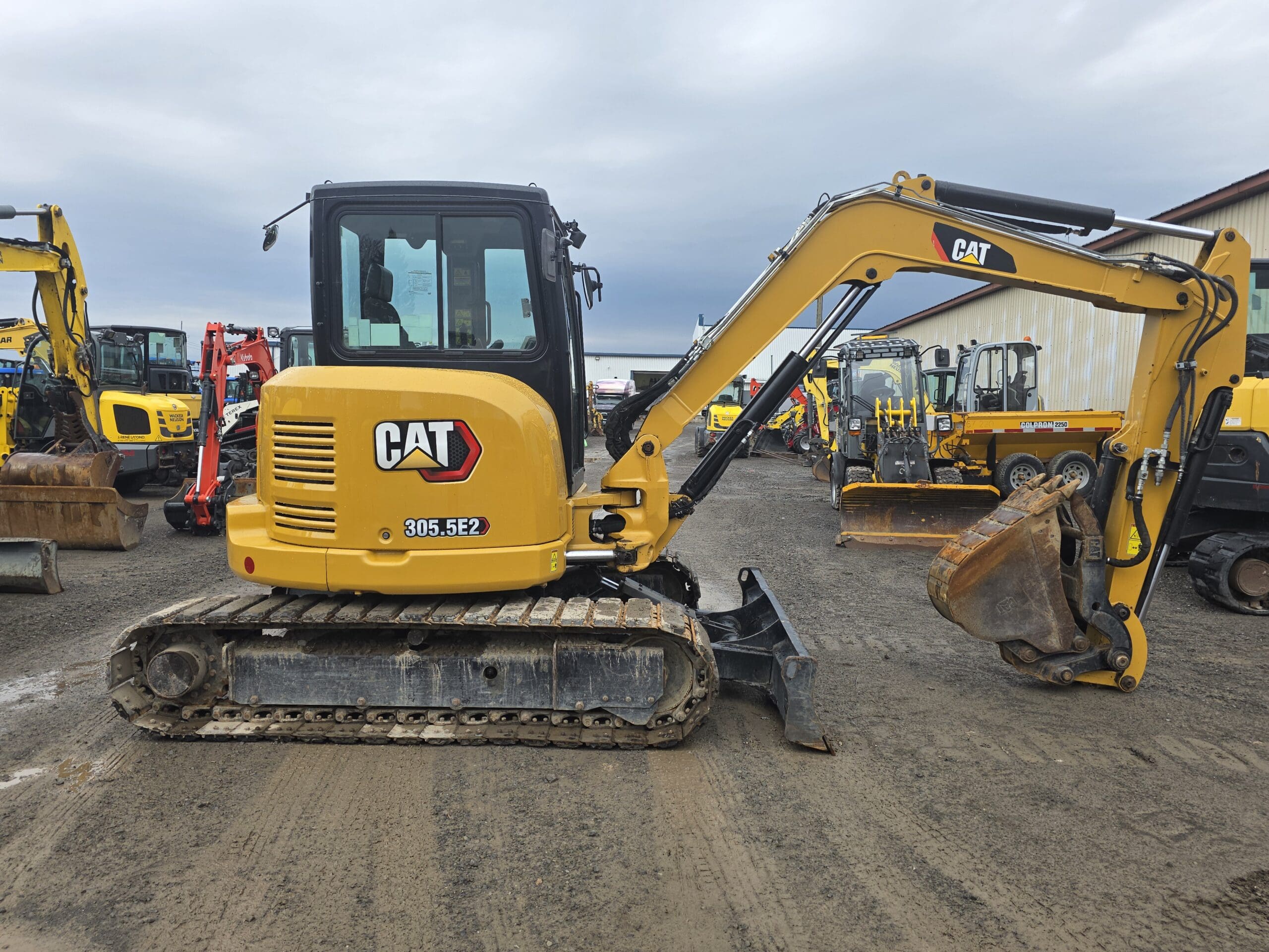 Une mini-pelle jaune CAT 305.5E2 est garée sur un terrain en terre parmi d'autres équipements de construction par une journée nuageuse.