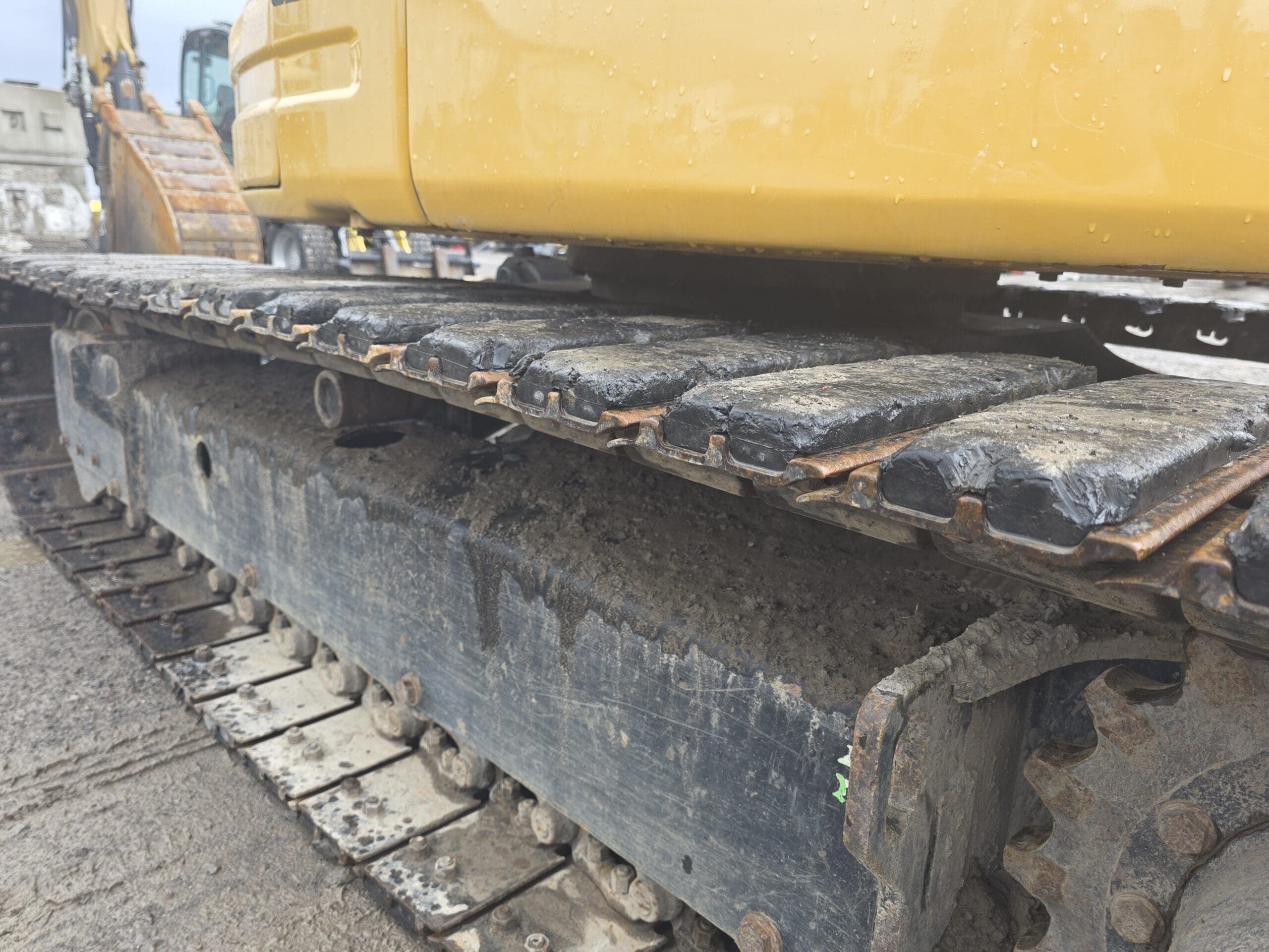 Gros plan des chenilles métalliques et du châssis d'une pelle sur chenilles, montrant des surfaces boueuses et usées, avec une partie de la carrosserie jaune visible au-dessus. La scène semble se dérouler sur un chantier de construction.