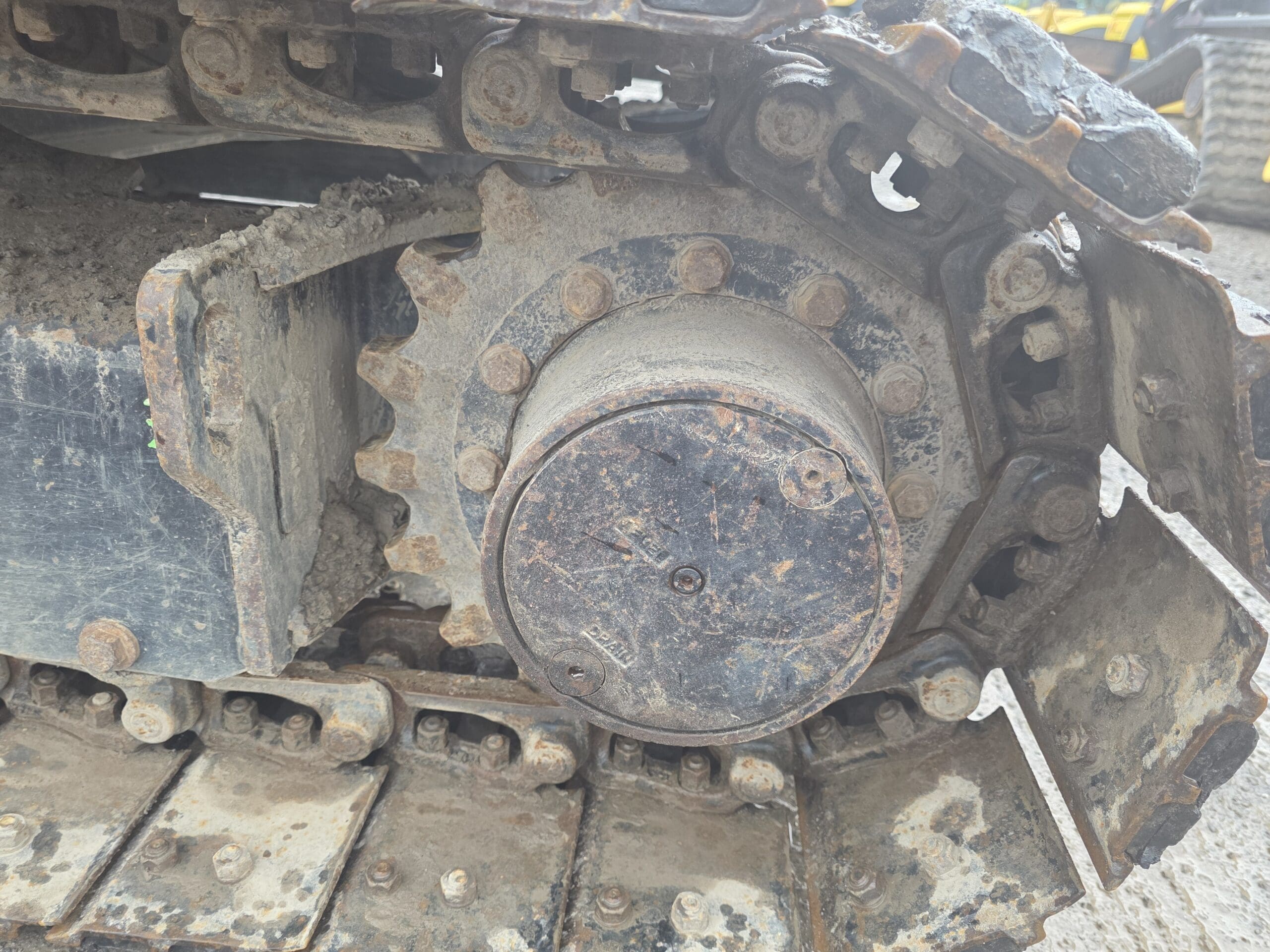 Vue rapprochée d'un pignon et d'une chenille en métal boueux sur un véhicule de construction, montrant des boulons et des maillons usés avec de la saleté et de la rouille sur les pièces de la machine.
