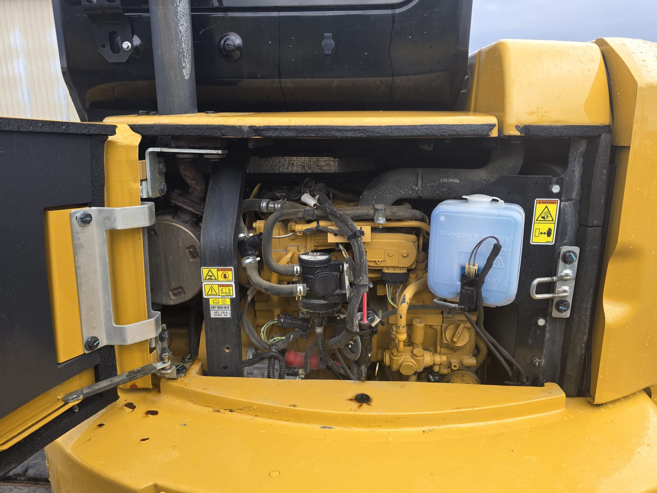 Gros plan du compartiment moteur d'un véhicule de construction jaune, montrant diverses pièces mécaniques, des étiquettes d'avertissement, des tuyaux et un récipient de liquide bleu translucide avec un bouchon, avec les panneaux métalliques environnants partiellement ouverts.
