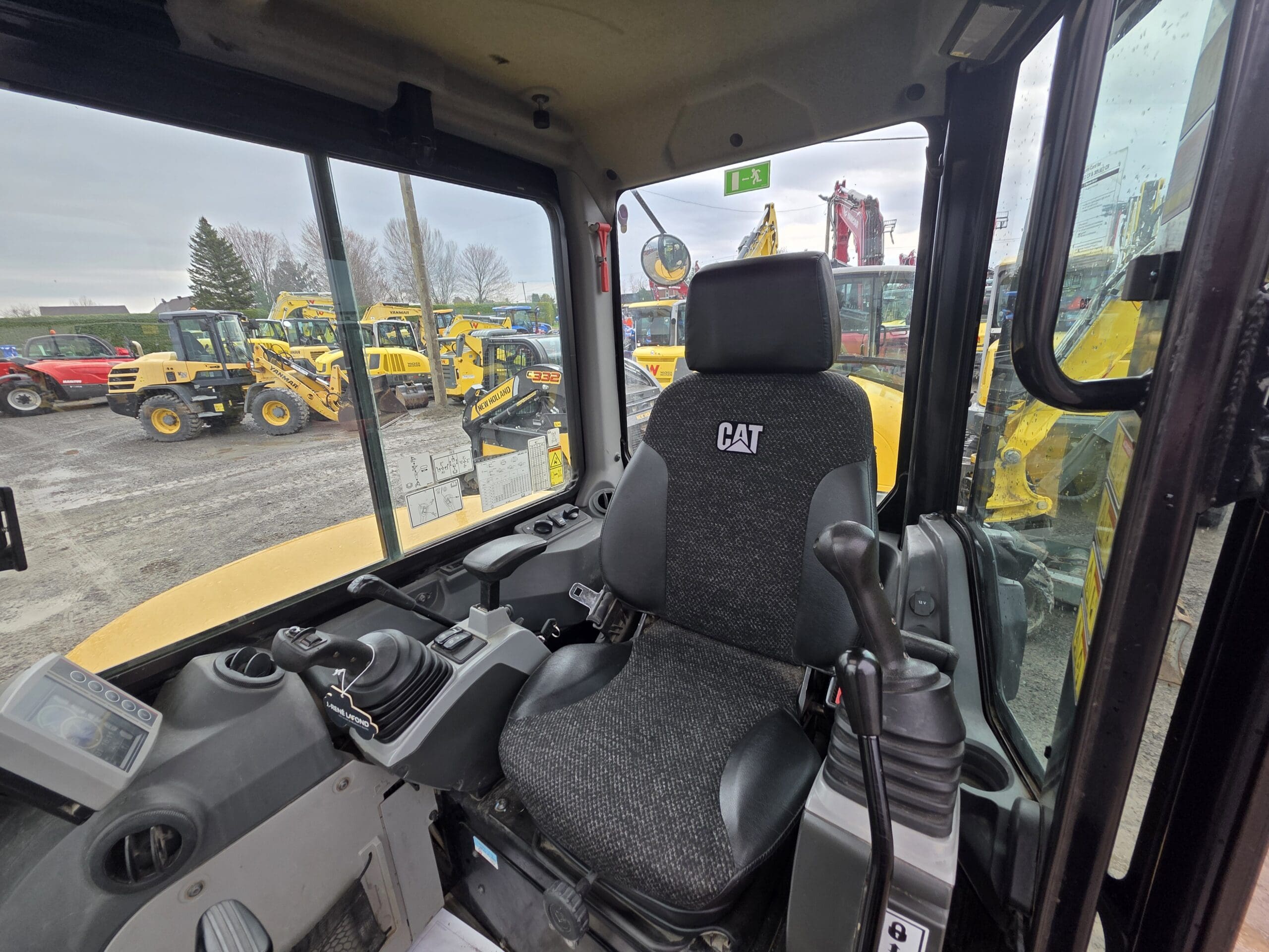 Vue de l'intérieur de la cabine d'un véhicule de construction CAT, montrant le siège du conducteur, les panneaux de commande et les leviers, avec diverses machines de construction jaunes visibles à l'extérieur à travers les fenêtres.