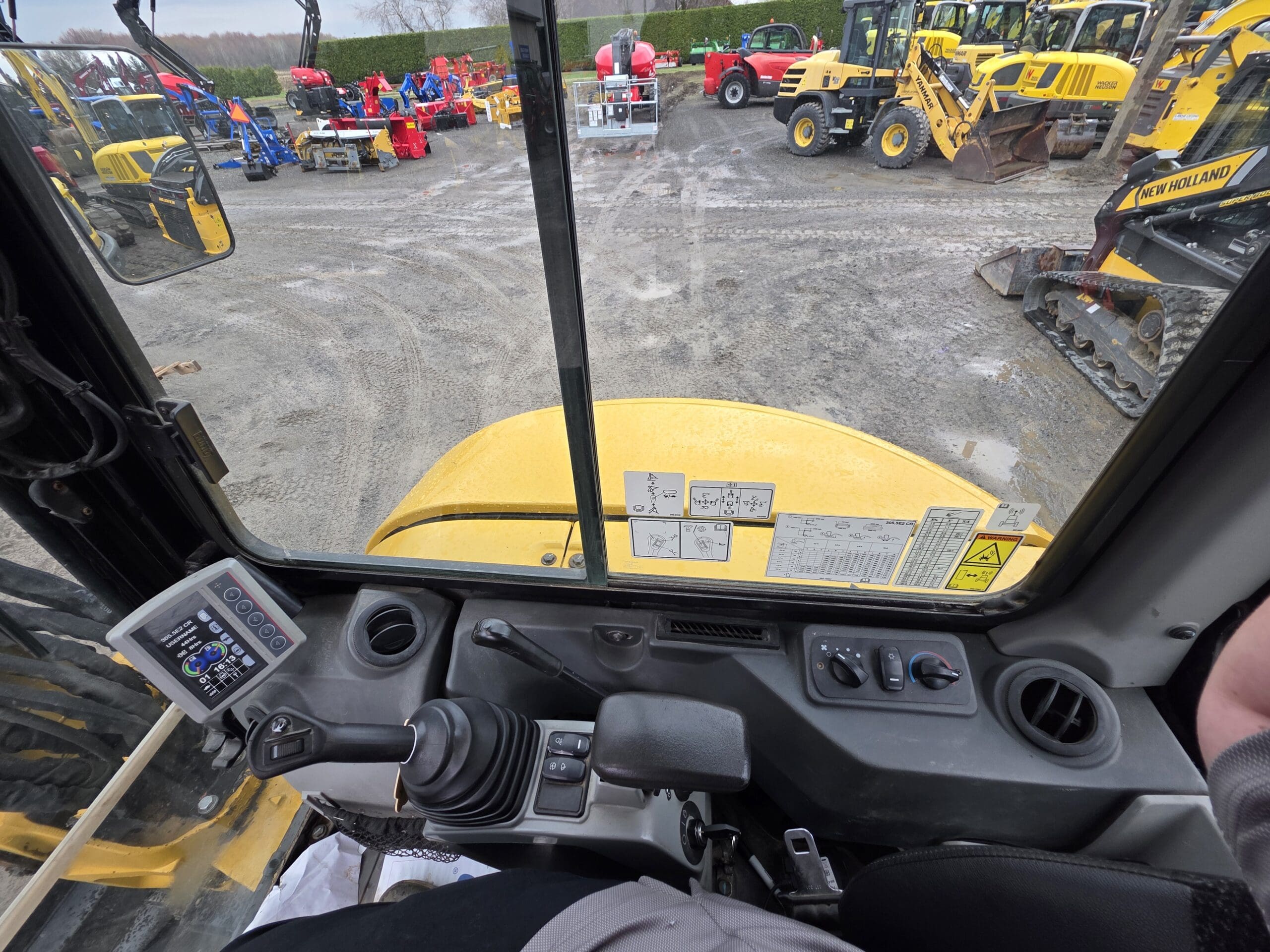 Vue de l'intérieur de la cabine d'un véhicule de construction, montrant des panneaux de commande, un joystick et des leviers, donnant sur un terrain avec divers équipements et machines de construction jaunes.