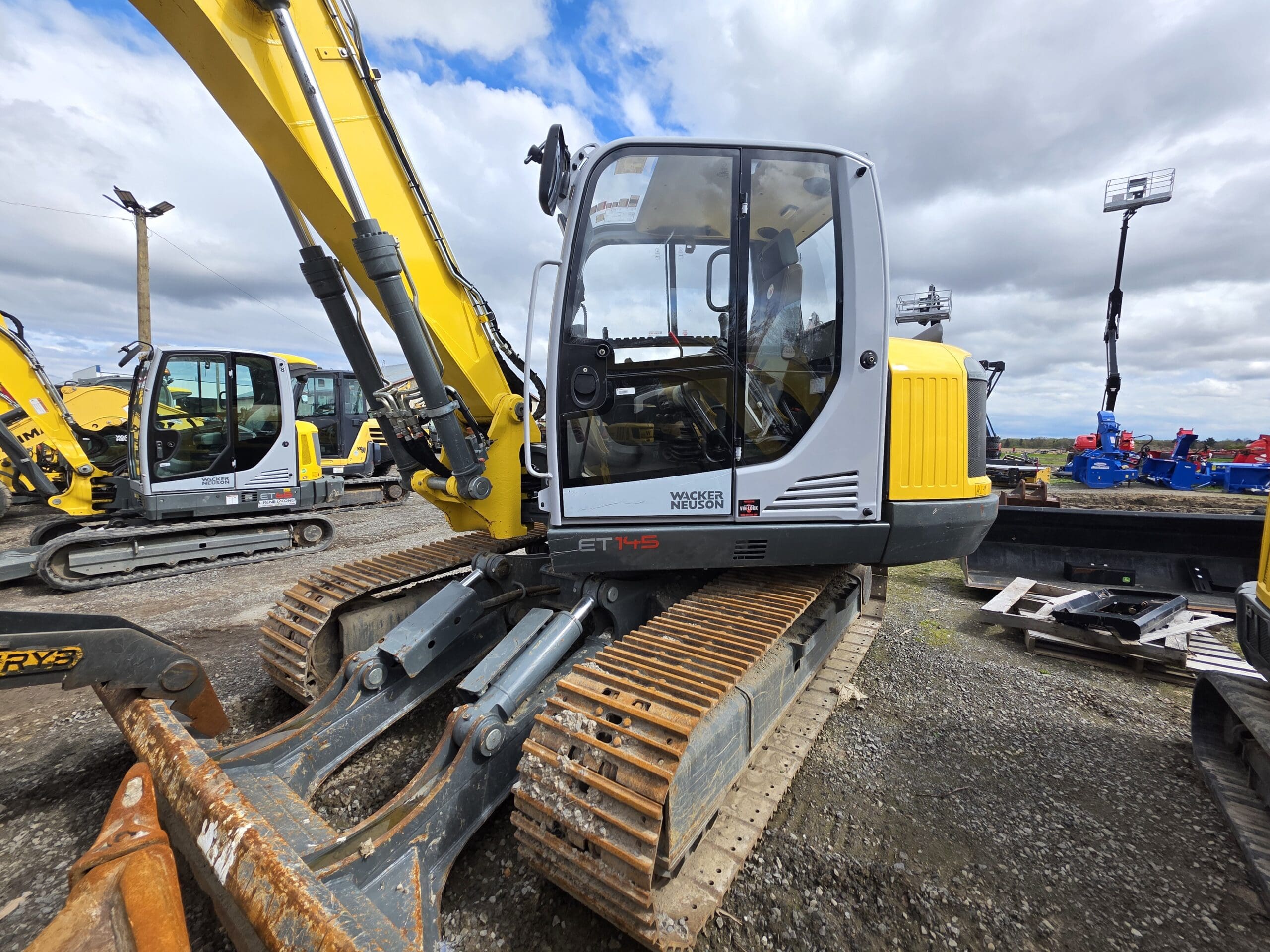 Une pelle compacte Wacker Neuson ET145 jaune et grise avec chenilles en caoutchouc est garée sur un terrain en gravier sous un ciel nuageux, entourée d'autres engins de chantier.