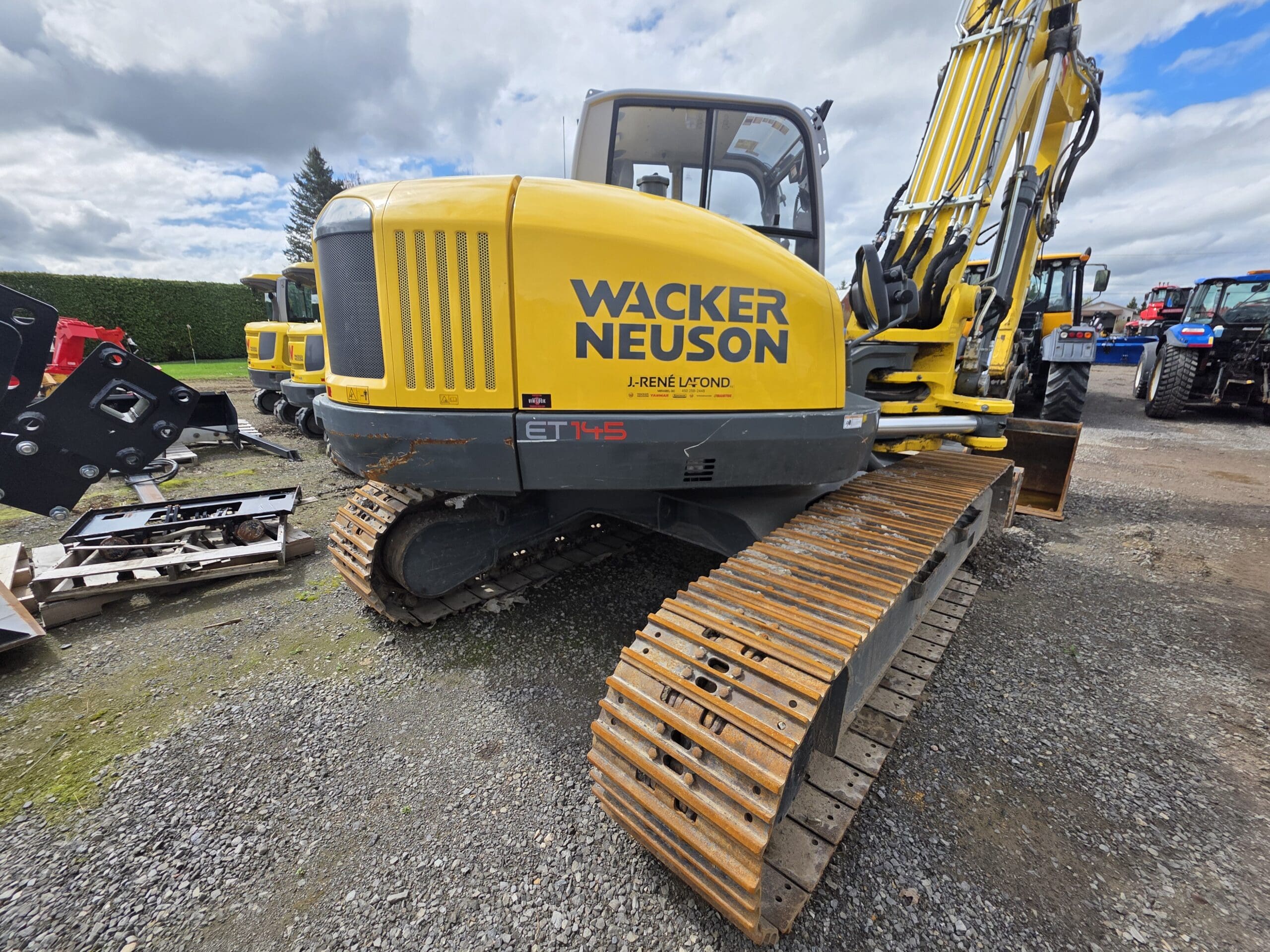 Une pelle sur chenilles jaune Wacker Neuson ET145 est garée sur du gravier, avec d'autres machines visibles en arrière-plan sous un ciel partiellement nuageux.