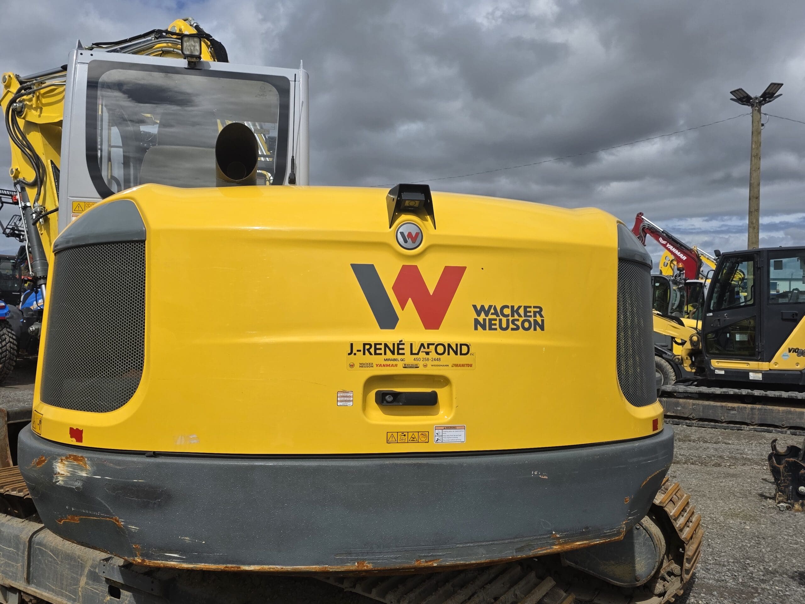 Une excavatrice jaune Wacker Neuson avec le logo et le nom « J. René Lafond » à l'arrière, stationnée sur un terrain en gravier avec d'autres véhicules de construction sous un ciel nuageux.