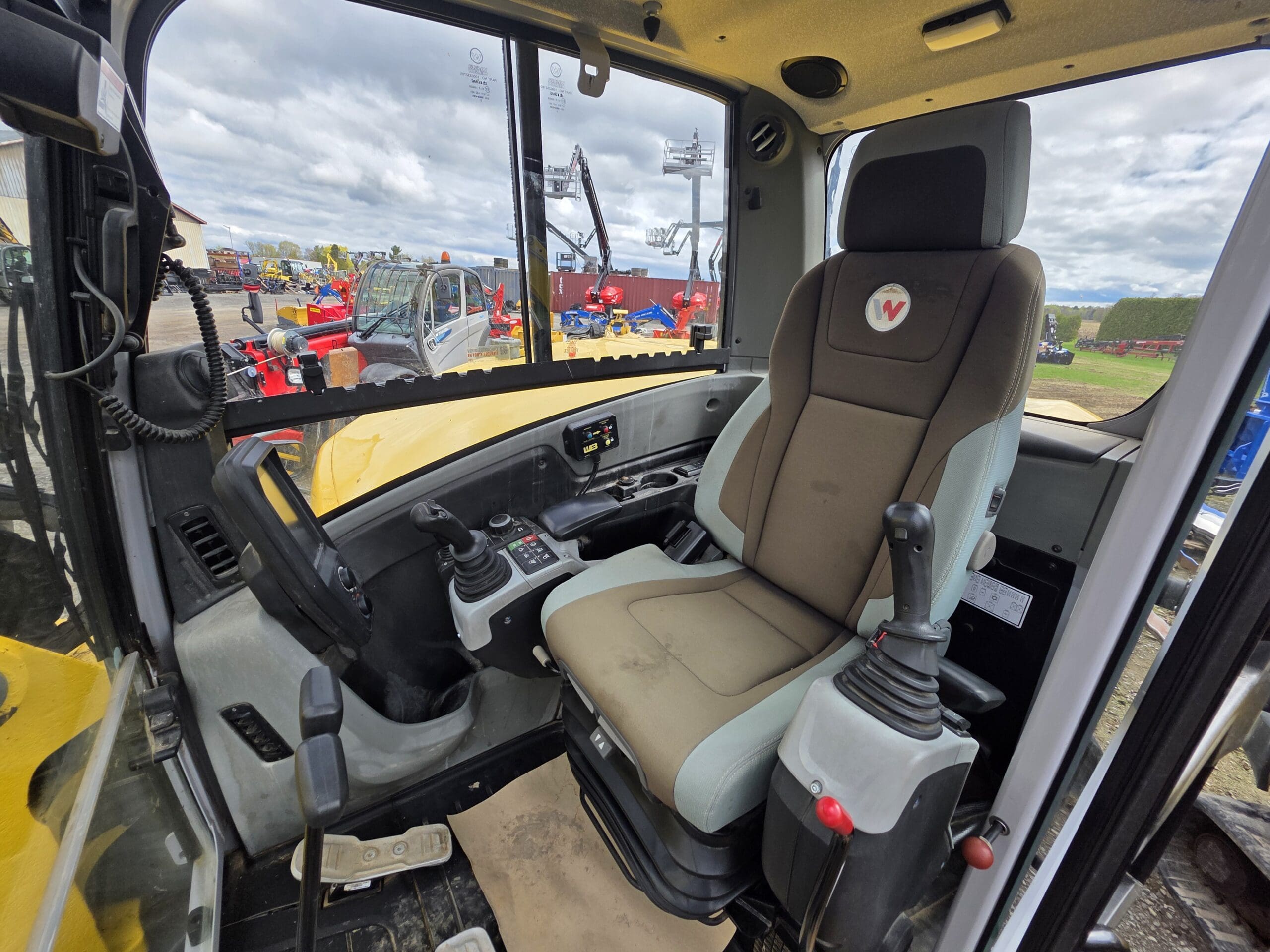 L'intérieur d'une cabine de véhicule de construction, montrant un siège conducteur rembourré, des joysticks de commande, des pédales et une vue à travers de grandes fenêtres sur les machines et équipements extérieurs par temps nuageux.