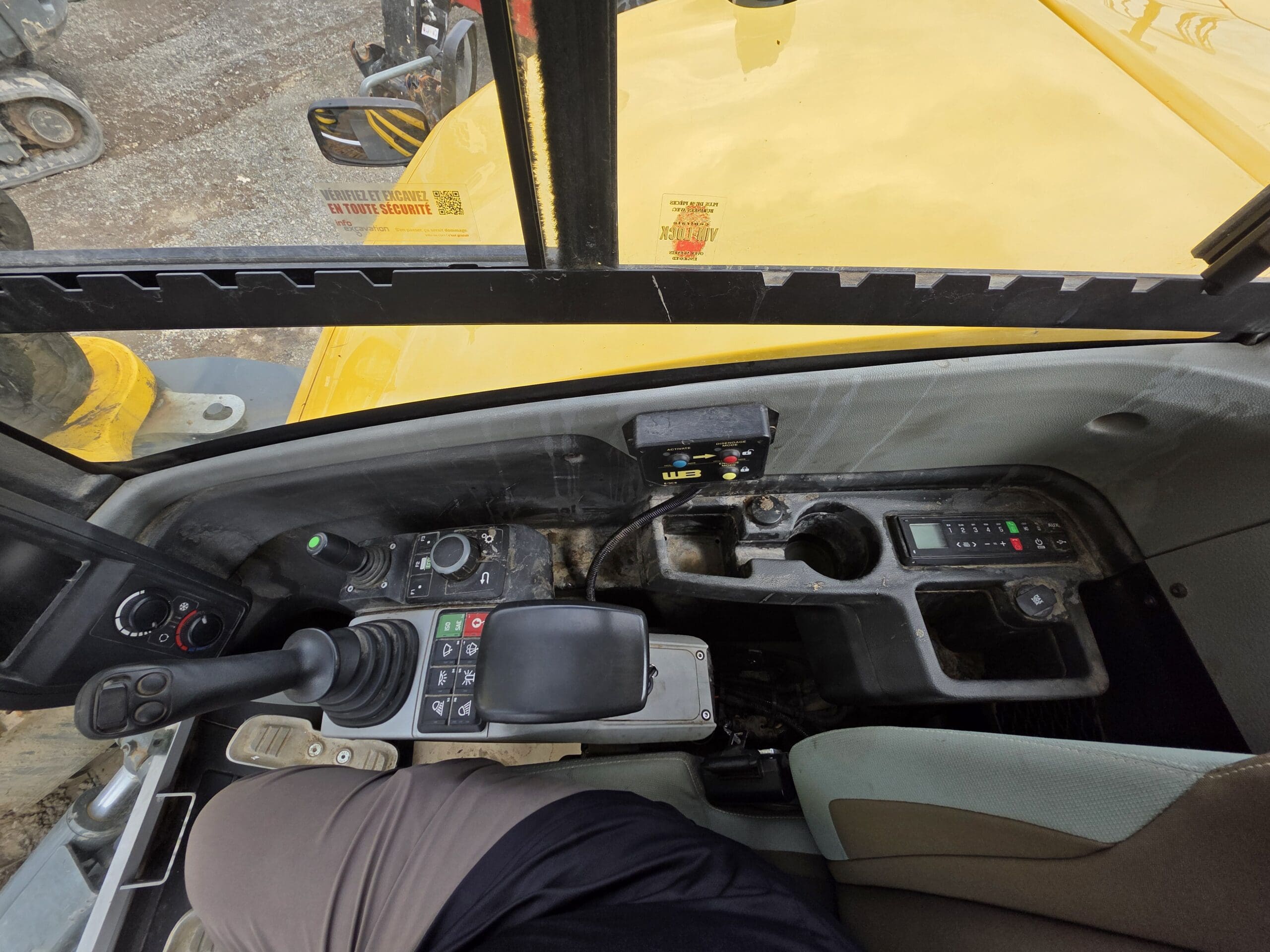 Vue de l'intérieur de la cabine d'un engin de chantier montrant les panneaux de commande, le joystick, l'accoudoir, les porte-gobelets et une partie du siège et de la jambe de l'opérateur. L'extérieur jaune du véhicule est visible par la vitre.