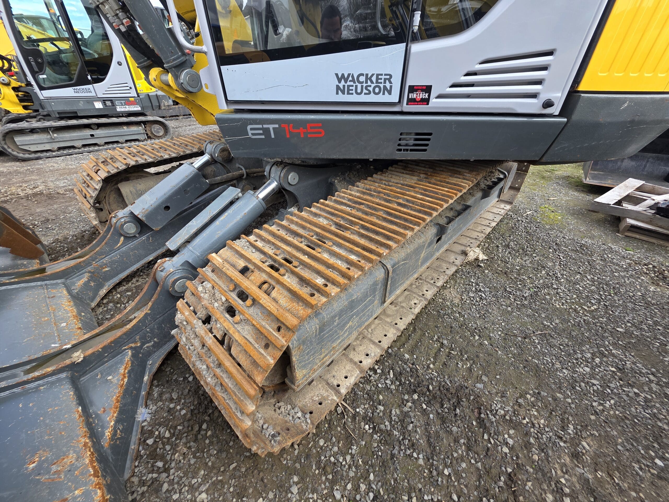 Gros plan des chenilles métalliques rouillées et du côté d'une excavatrice Wacker Neuson ET145 sur une surface de gravier, avec un autre véhicule de construction visible en arrière-plan.