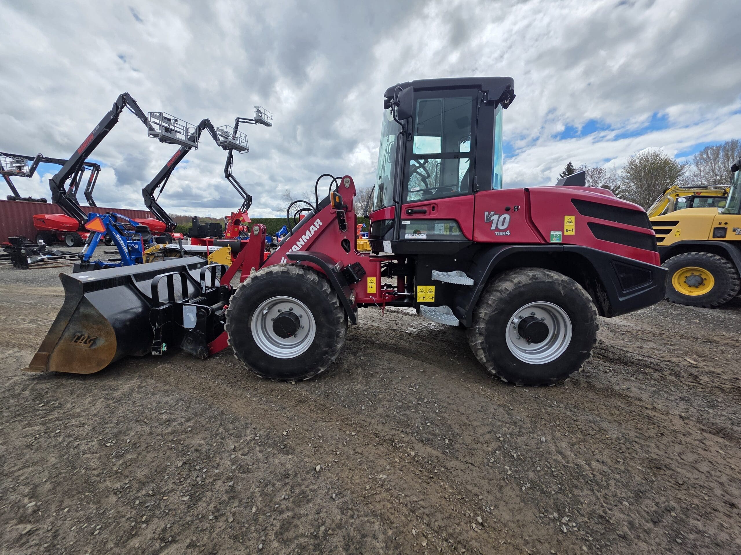 Une chargeuse sur pneus Yanmar V100 rouge avec de gros pneus est garée sur du gravier dans un parc d'équipements de construction, avec d'autres machines visibles en arrière-plan sous un ciel nuageux.