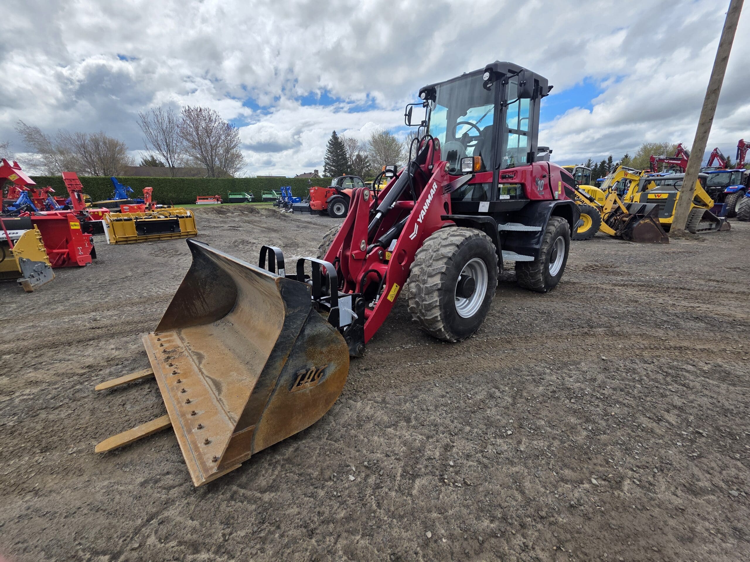 Une chargeuse frontale rouge avec un grand godet est garée sur un terrain en terre sous un ciel nuageux, entourée d'autres machines de construction et agricoles.