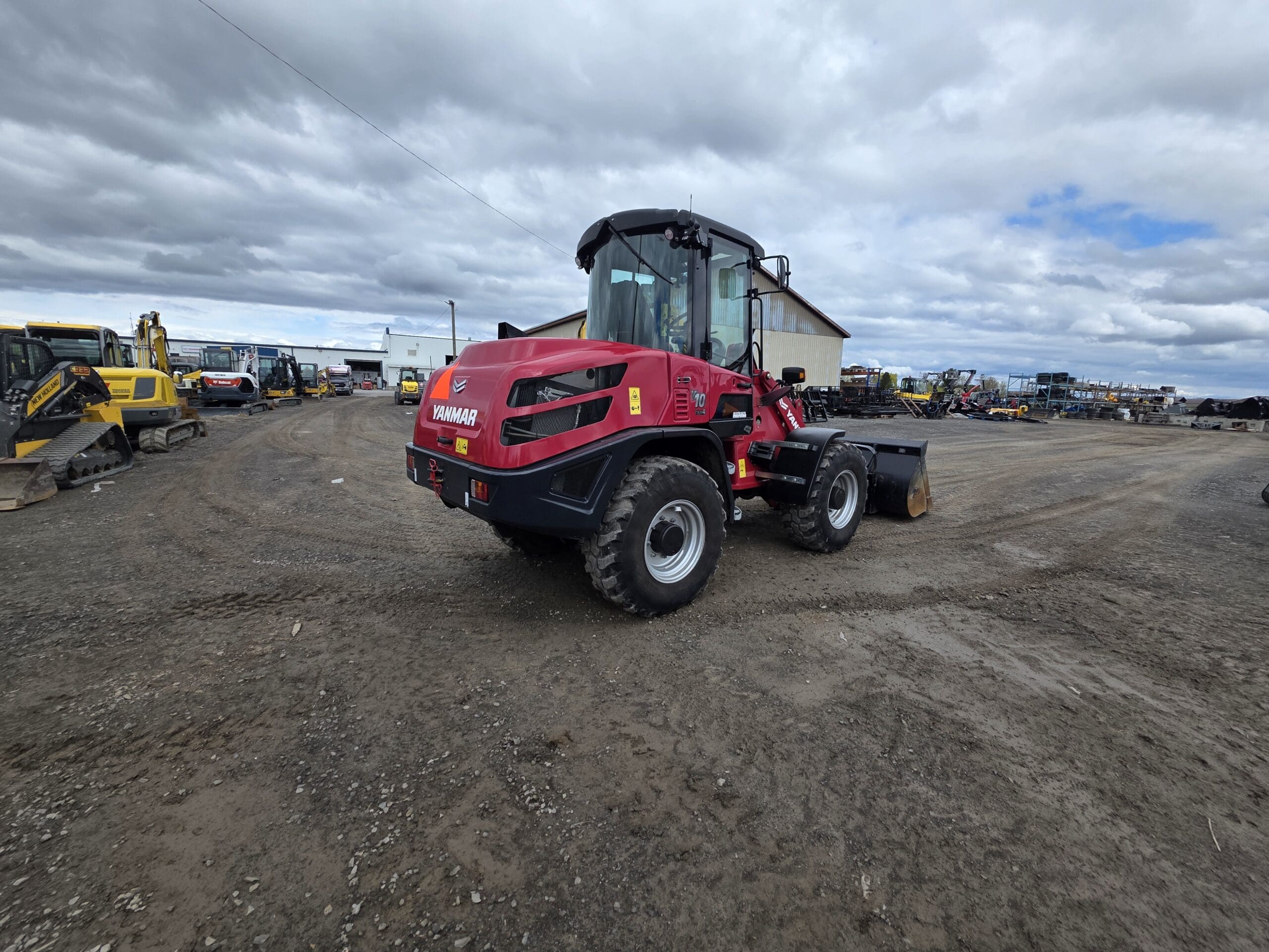 Un véhicule de construction Yanmar rouge est garé sur un terrain en terre sous un ciel nuageux, entouré d'autres machines lourdes et d'équipements industriels.
