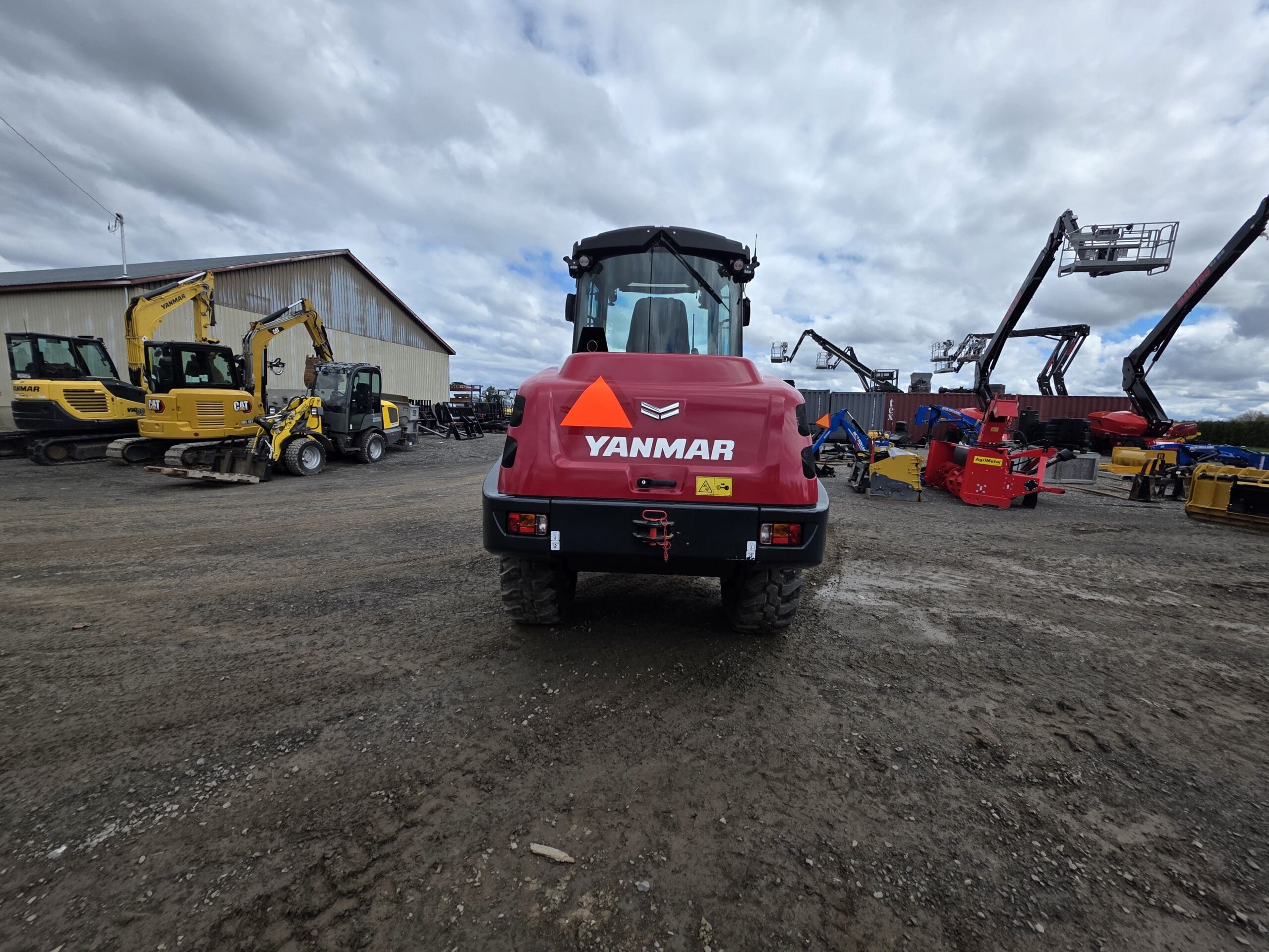 Un véhicule de construction Yanmar rouge avec un panneau orange triangulaire est garé sur un terrain en gravier, entouré de divers équipements de construction sous un ciel nuageux.