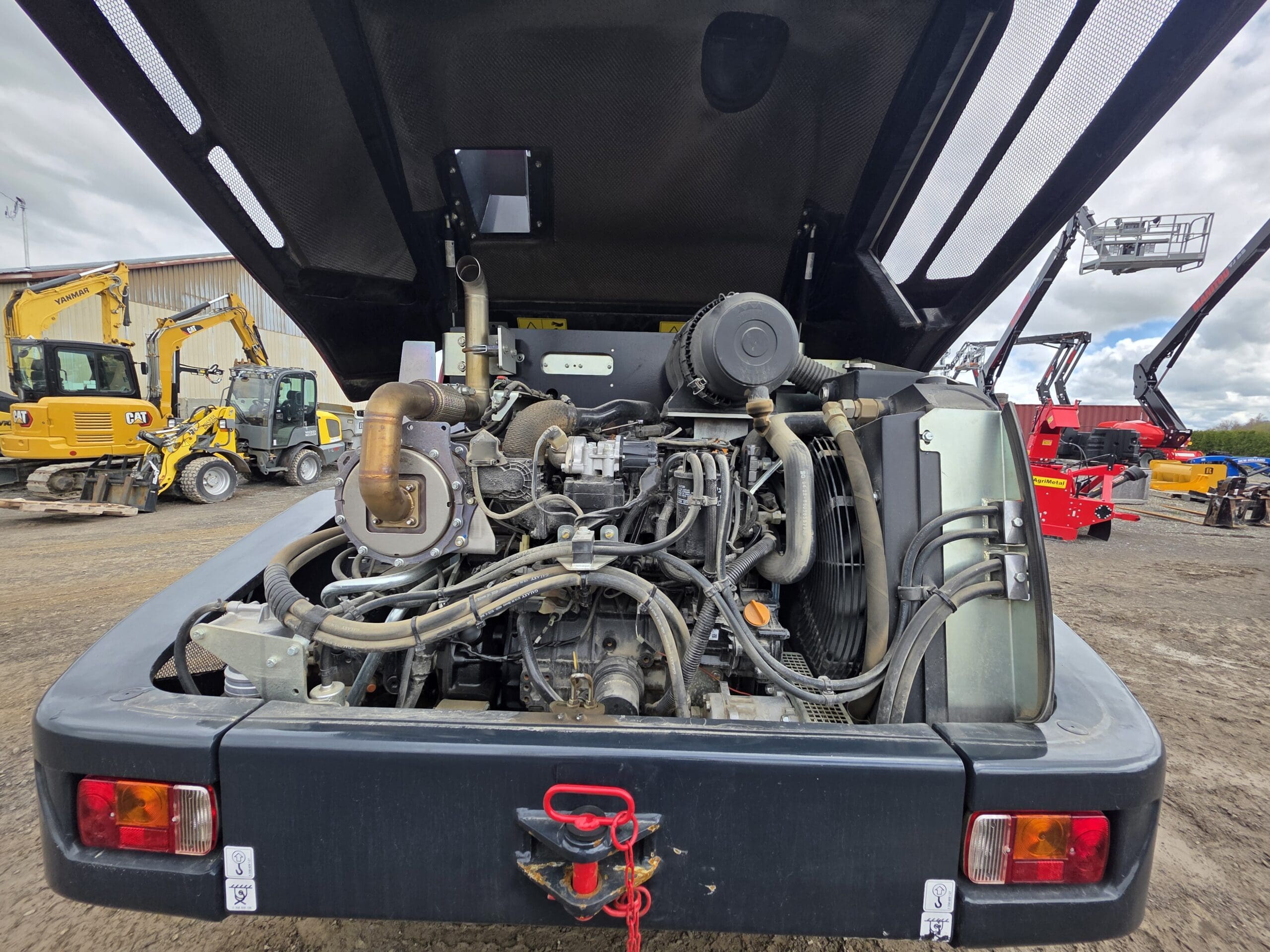 Vue rapprochée du moteur exposé et des composants mécaniques à l'arrière d'un véhicule de construction, avec diverses machines et équipements visibles en arrière-plan sur un chantier.
