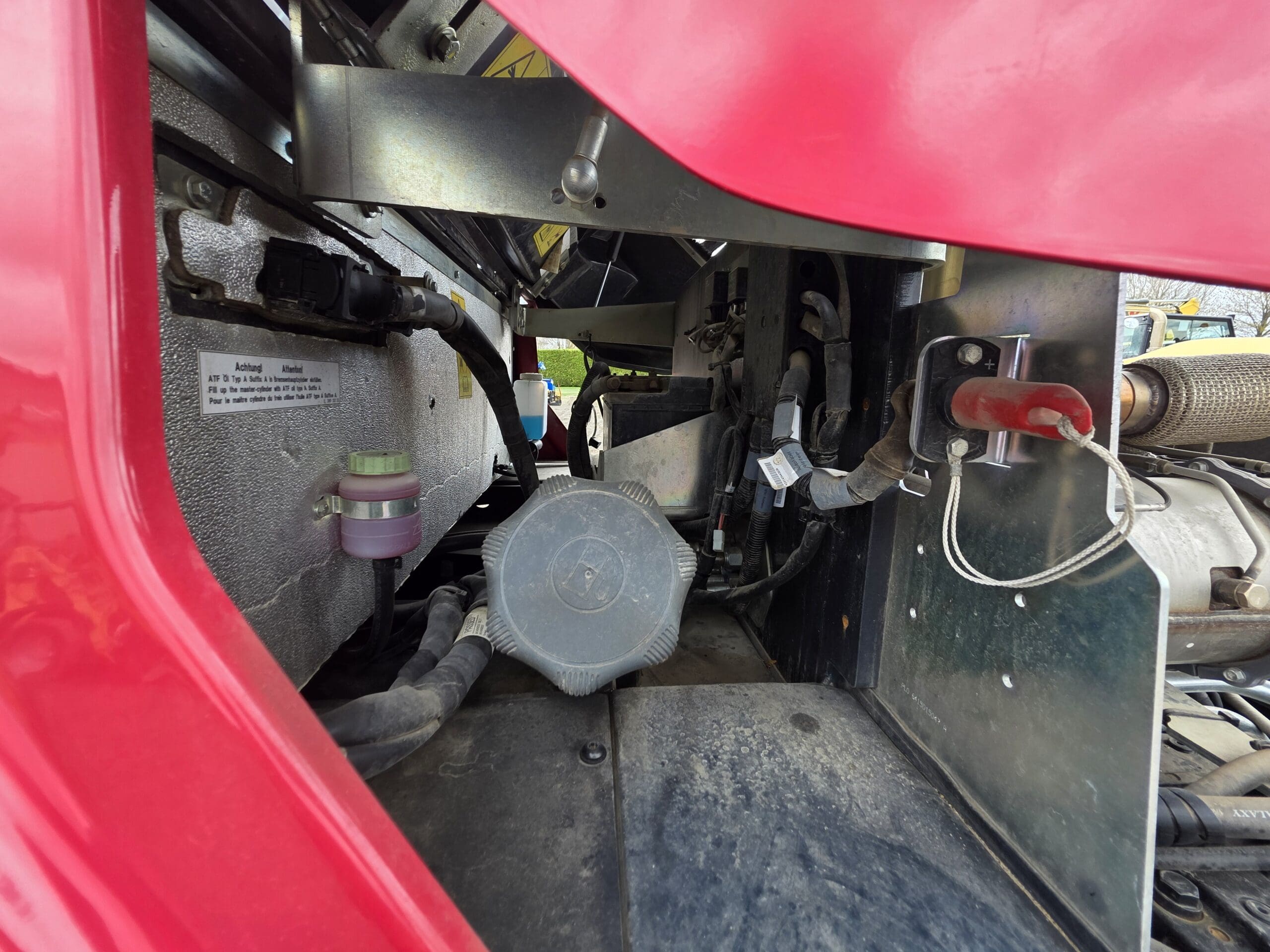Vue rapprochée du compartiment moteur d’un camion, montrant divers composants mécaniques, des tuyaux et un réservoir de liquide de refroidissement sous un capot rouge partiellement surélevé.