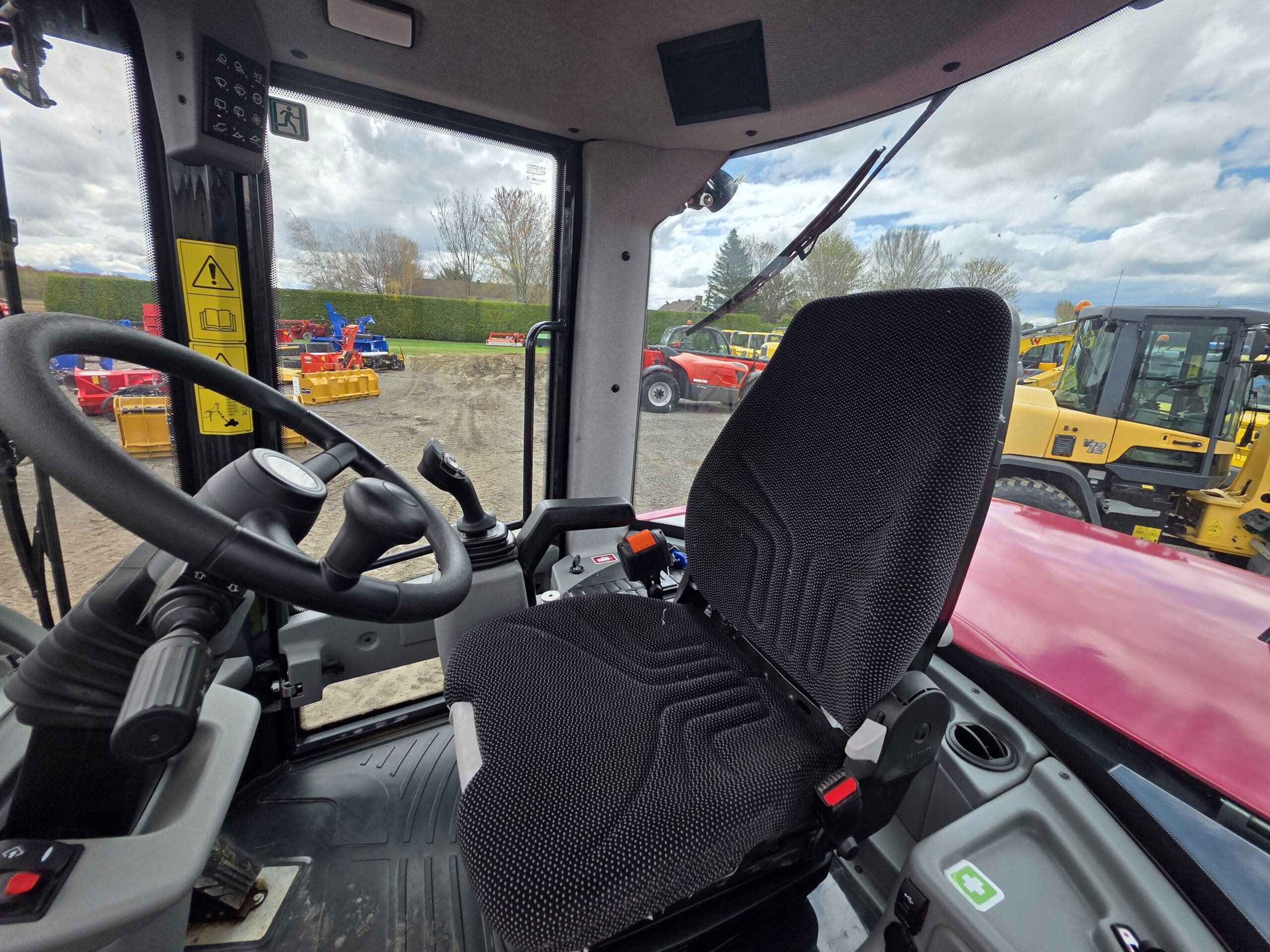 Vue intérieure d'une cabine de tracteur montrant un siège conducteur noir, un volant et des commandes, avec des véhicules de construction et agricoles visibles à l'extérieur à travers de grandes fenêtres par temps nuageux.
