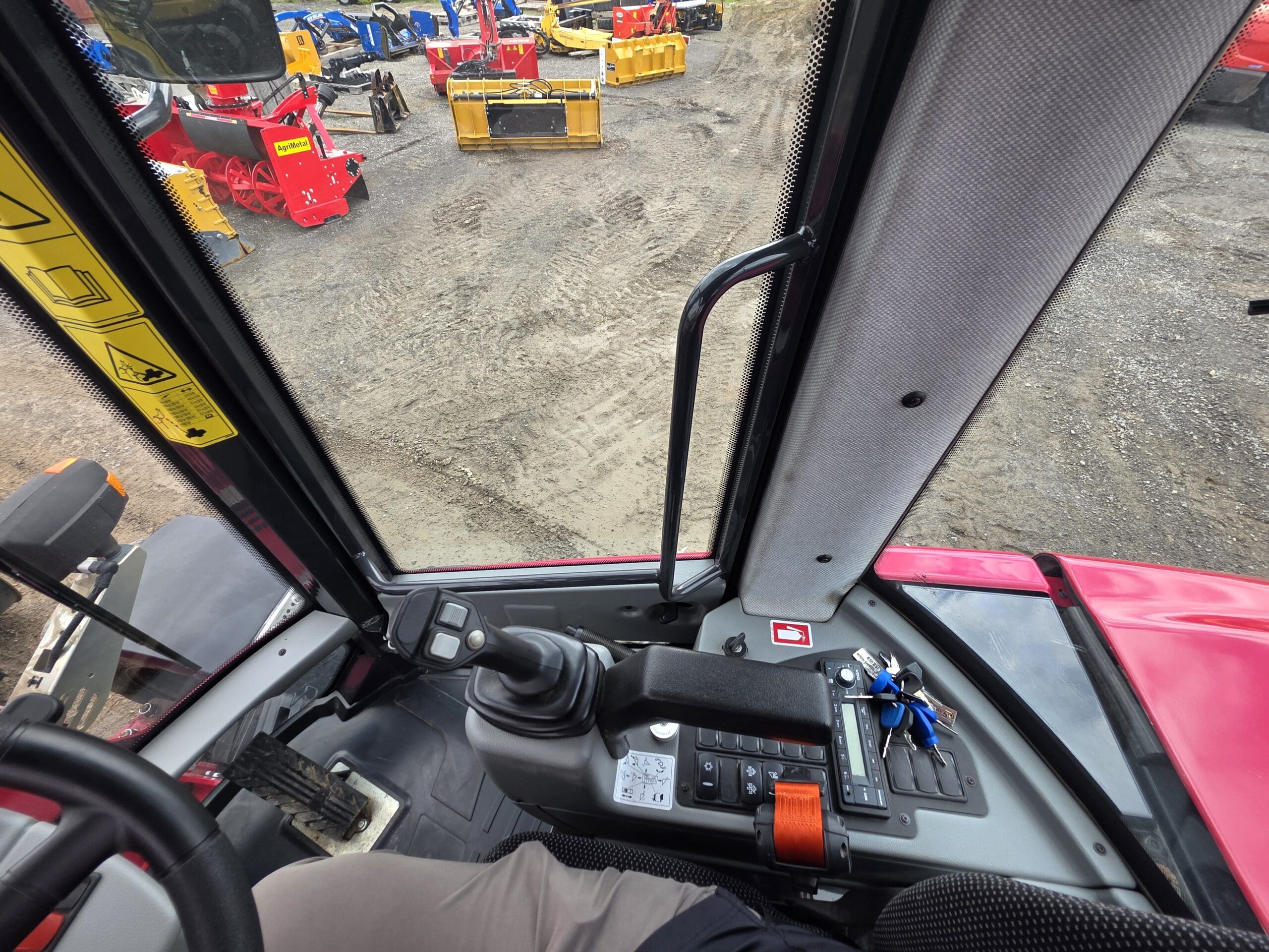 Vue de l'intérieur d'une cabine de tracteur montrant des leviers de commande, des boutons, un joystick et un jeu de clés, avec des équipements de construction visibles à l'extérieur sur un terrain en gravier.