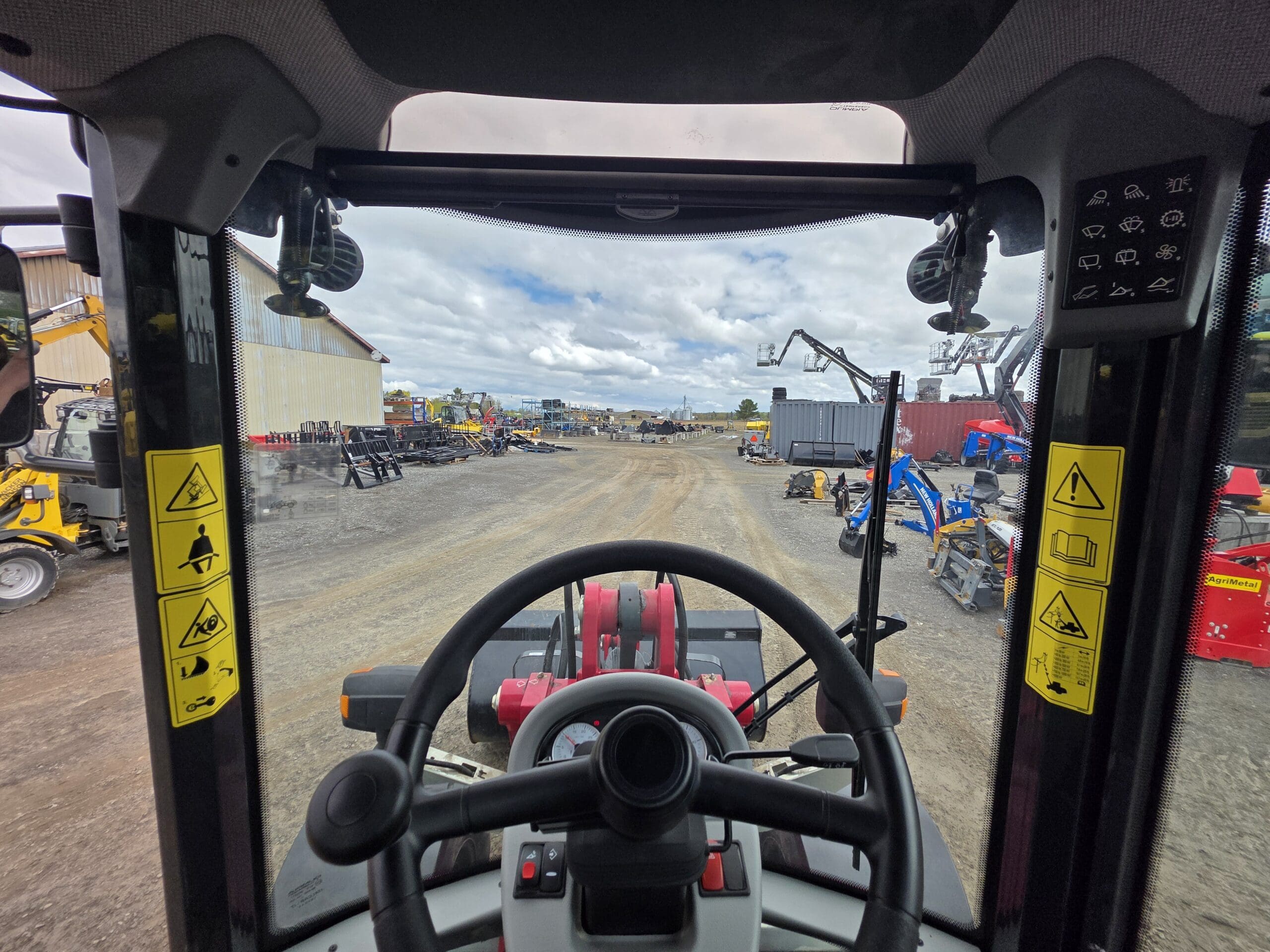 Vue depuis la cabine d'un tracteur donnant sur un chantier de construction, avec des machines et équipements stationnés de part et d'autre d'un chemin de terre sous un ciel nuageux. Des autocollants d'avertissement sont visibles sur le châssis de la cabine.