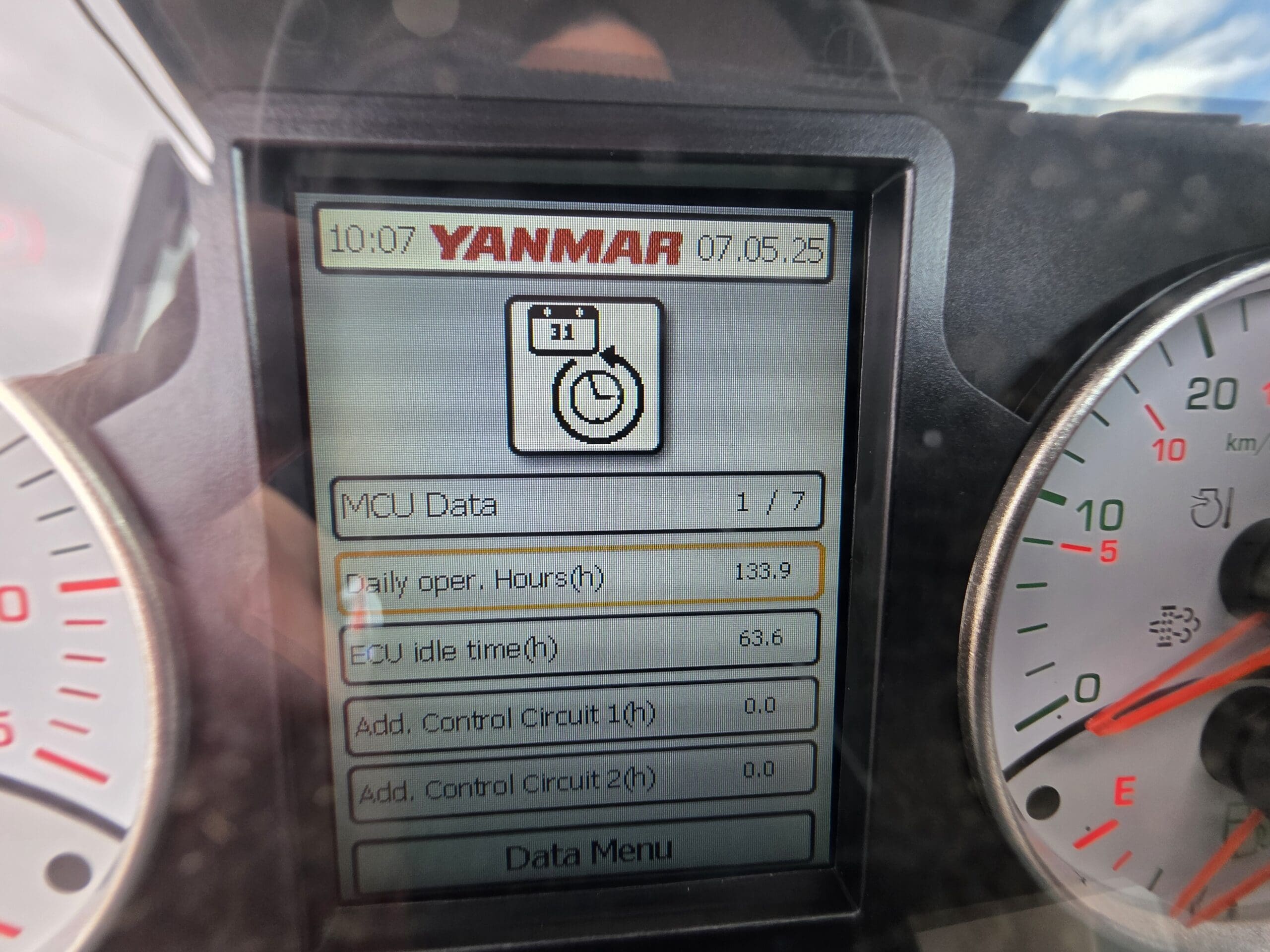 Gros plan d'un écran numérique sur une machine Yanmar affichant des données opérationnelles, notamment les heures de fonctionnement quotidiennes (133,9) et le temps d'inactivité de l'ECU (63,6 heures), avec deux jauges analogiques partiellement visibles de chaque côté.
