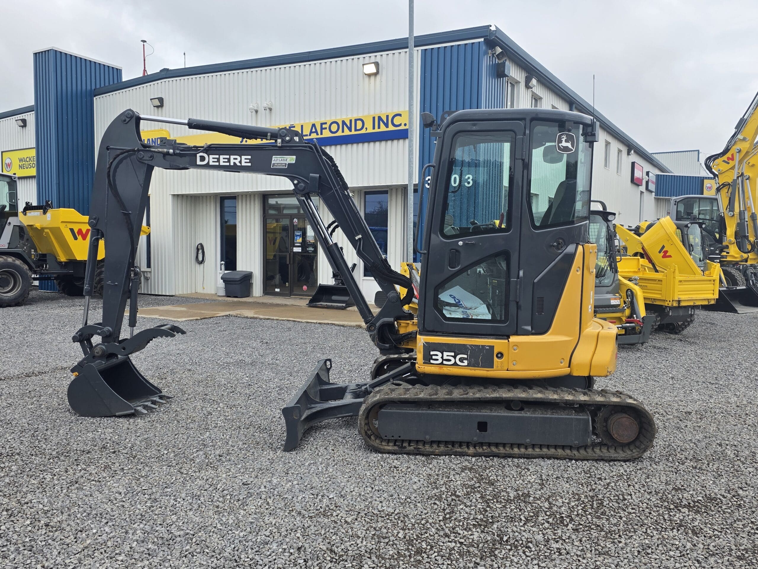 Une pelle compacte John Deere 35G, dotée d'un bras noir et d'une benne jaune, est garée sur du gravier devant un bâtiment industriel bleu et blanc. D'autres engins de chantier sont visibles en arrière-plan.