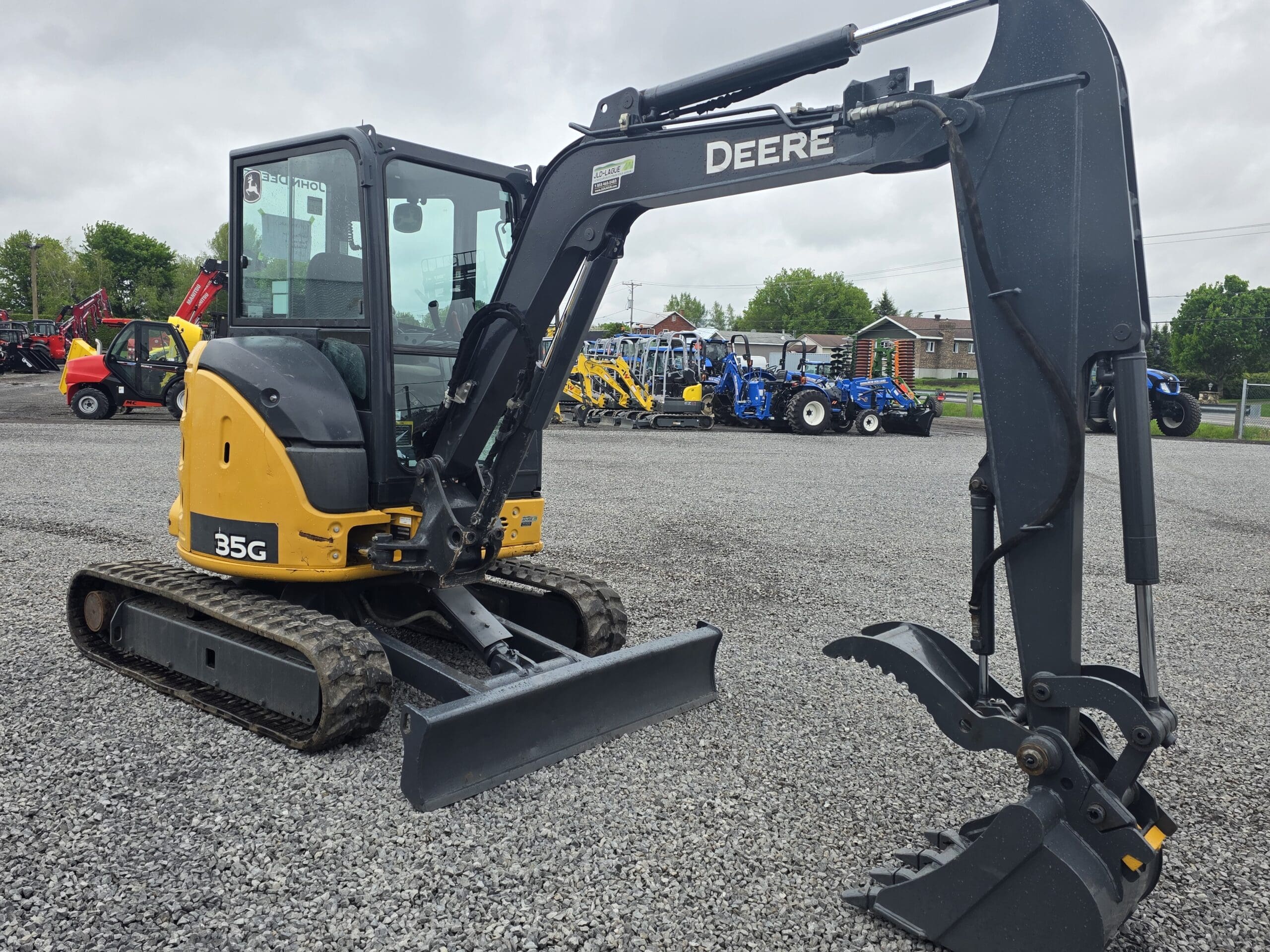 Une mini-pelle John Deere 35G jaune et noire avec un grappin est garée sur du gravier, entourée d'autres machines par une journée nuageuse.
