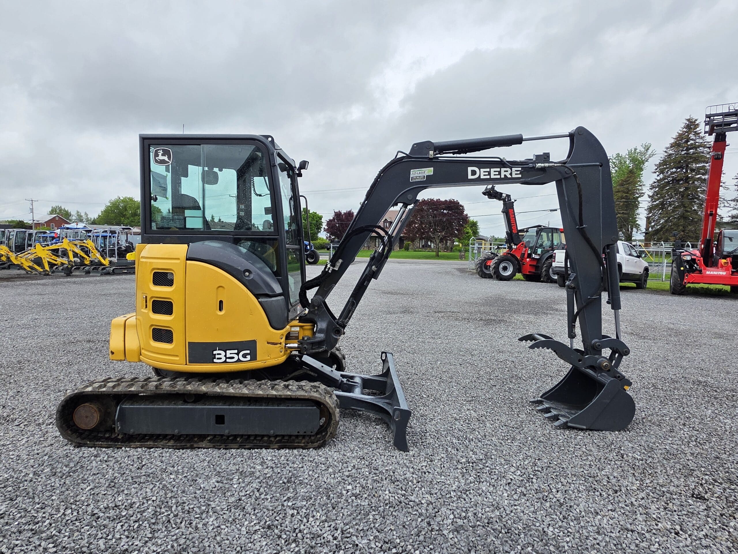 Une excavatrice compacte John Deere 35G jaune et noire avec un accessoire à griffes est garée sur un terrain en gravier avec d'autres équipements lourds en arrière-plan par une journée nuageuse.