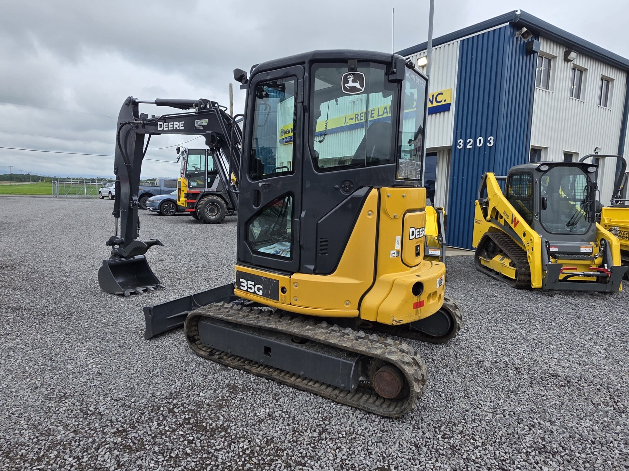 Une mini-pelle John Deere 35G jaune et noire avec une cabine est garée sur du gravier à côté d'un bâtiment bleu et blanc, avec d'autres équipements de construction visibles en arrière-plan.