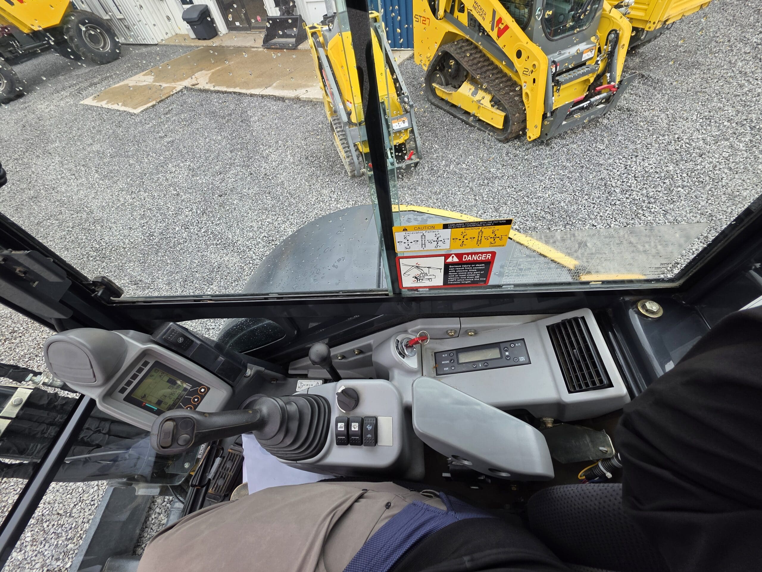 Vue de l'intérieur de la cabine d'un engin de chantier, montrant le panneau de commande, le joystick et l'écran. À l'extérieur, un engin de chantier jaune est garé sur du gravier près d'un bâtiment. Des étiquettes d'avertissement sont visibles sur la vitre.