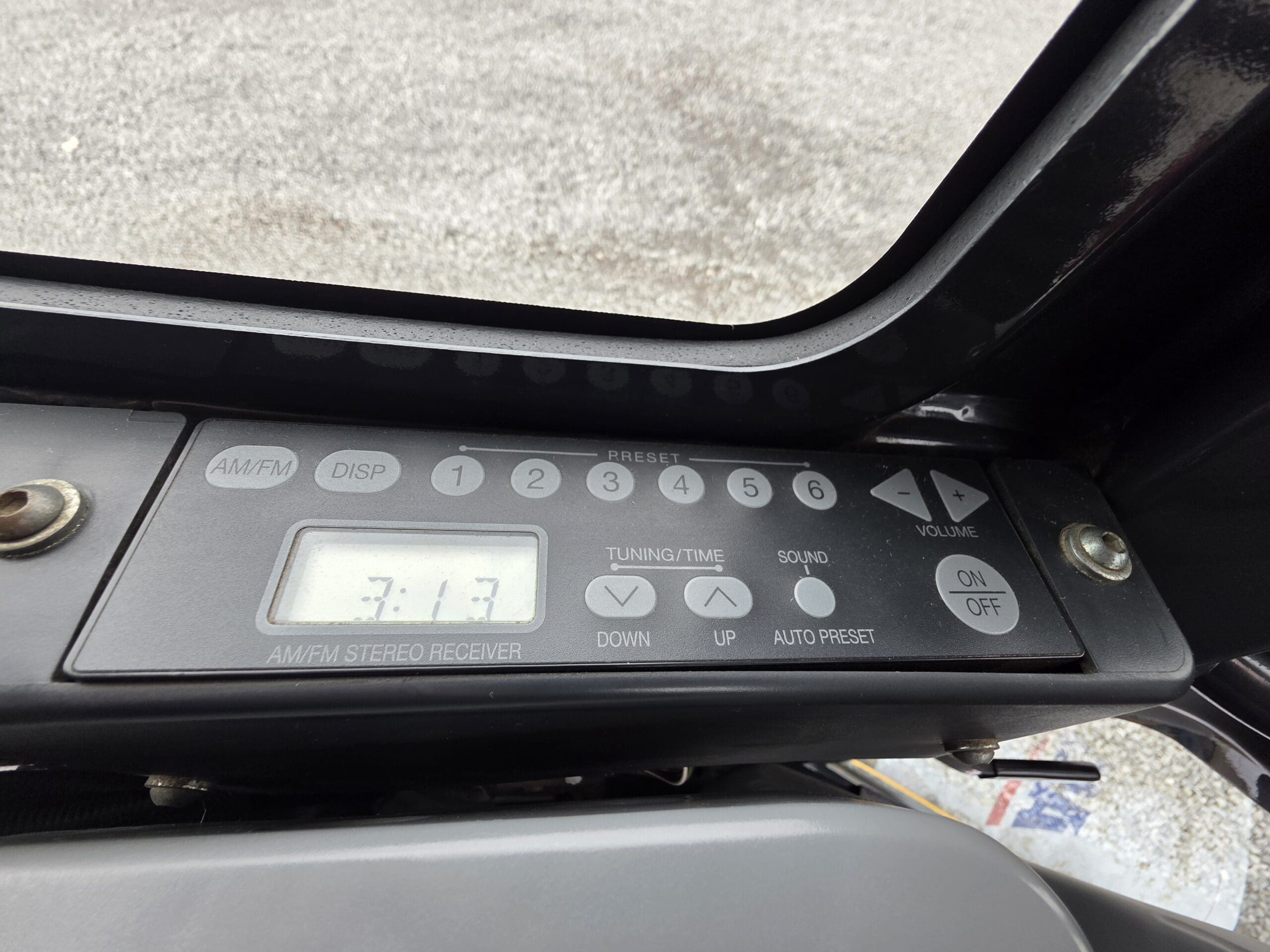 Gros plan sur le panneau de commande d'un autoradio avec boutons de préréglage, un écran indiquant « 7:17 », des commandes de réglage et de volume, et un bouton « Marche/Arrêt ». L'arrière-plan montre une surface de gravier à travers une fenêtre.