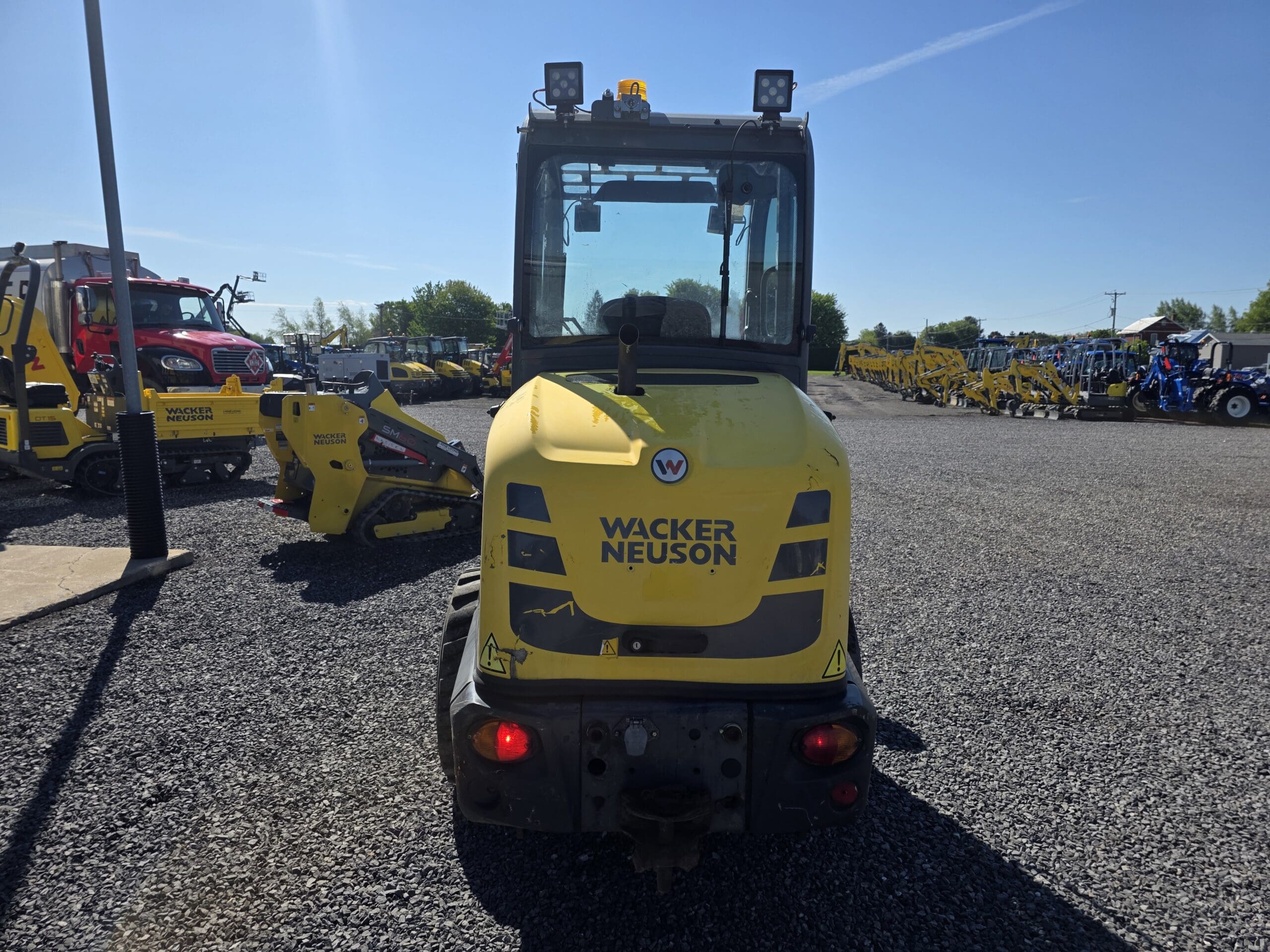 Un véhicule de construction jaune Wacker Neuson est garé sur du gravier dans un parking avec d'autres machines, vu de l'arrière sous un ciel bleu clair.