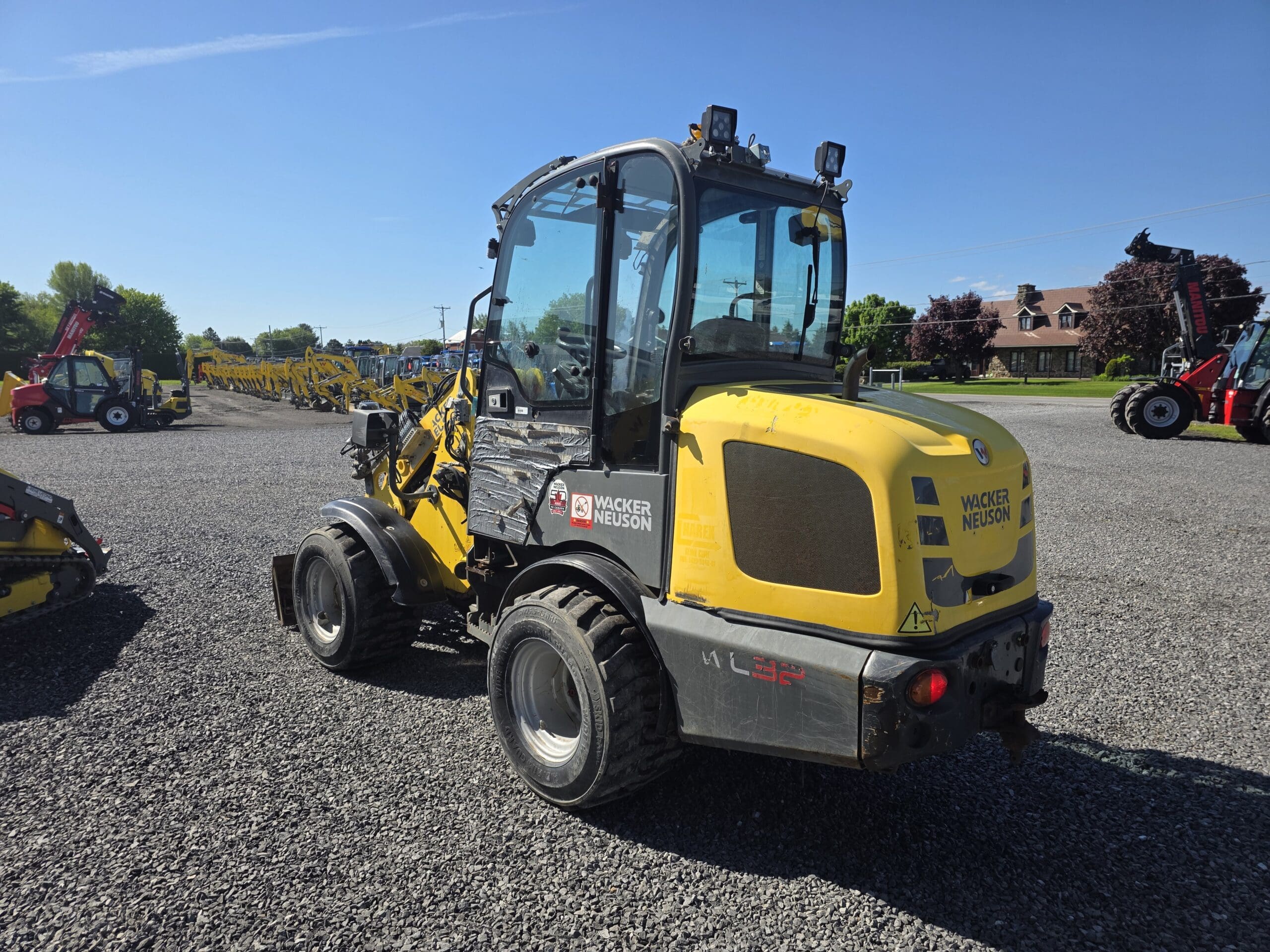 Une chargeuse sur pneus jaune Wacker Neuson est garée sur un terrain en gravier sous un ciel ensoleillé, avec divers équipements de construction et un bâtiment visibles en arrière-plan.