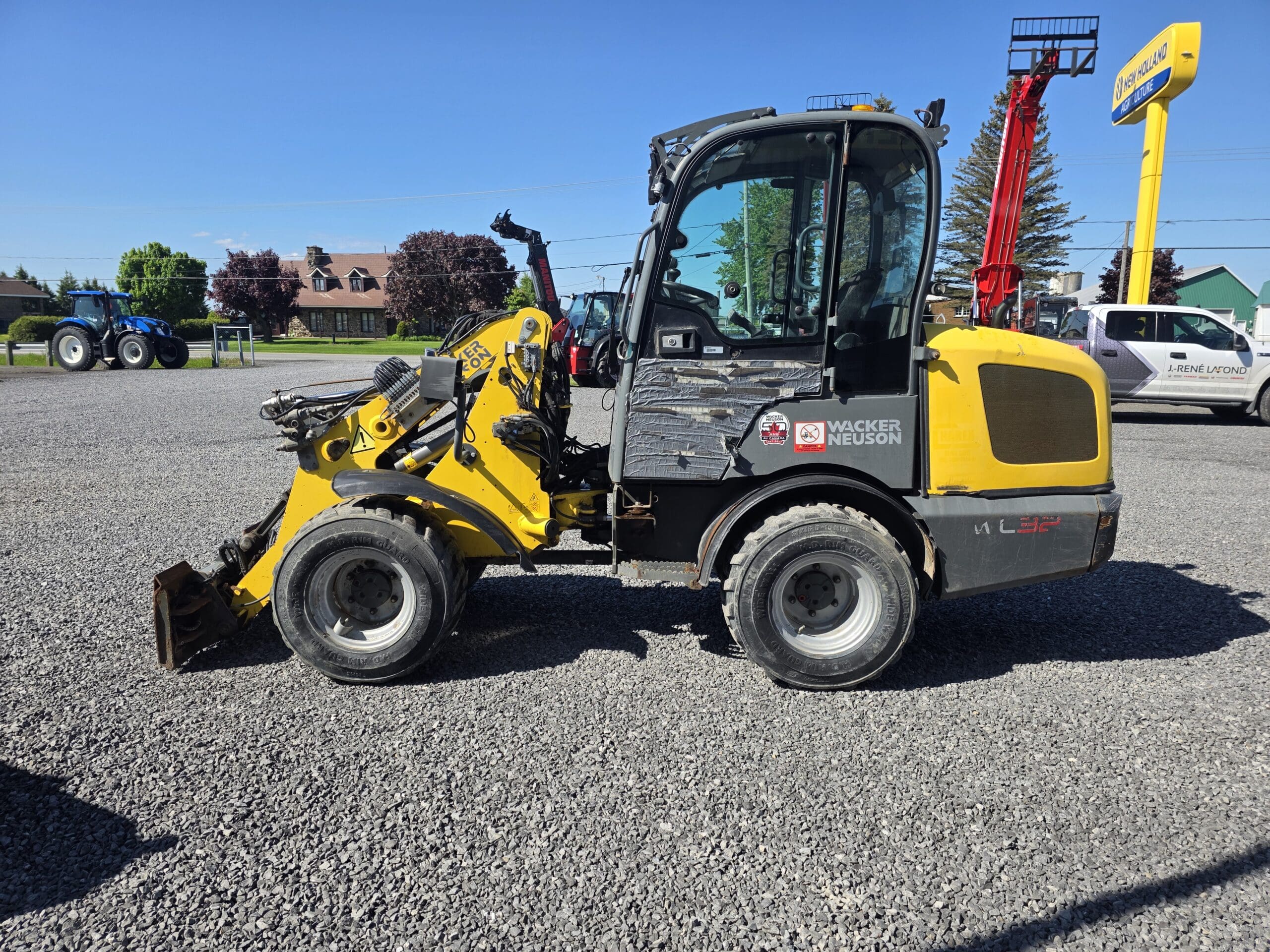 Une chargeuse sur pneus Wacker Neuson jaune et noire est garée sur un terrain en gravier sous un ciel bleu clair, avec d'autres machines, bâtiments et arbres visibles en arrière-plan.