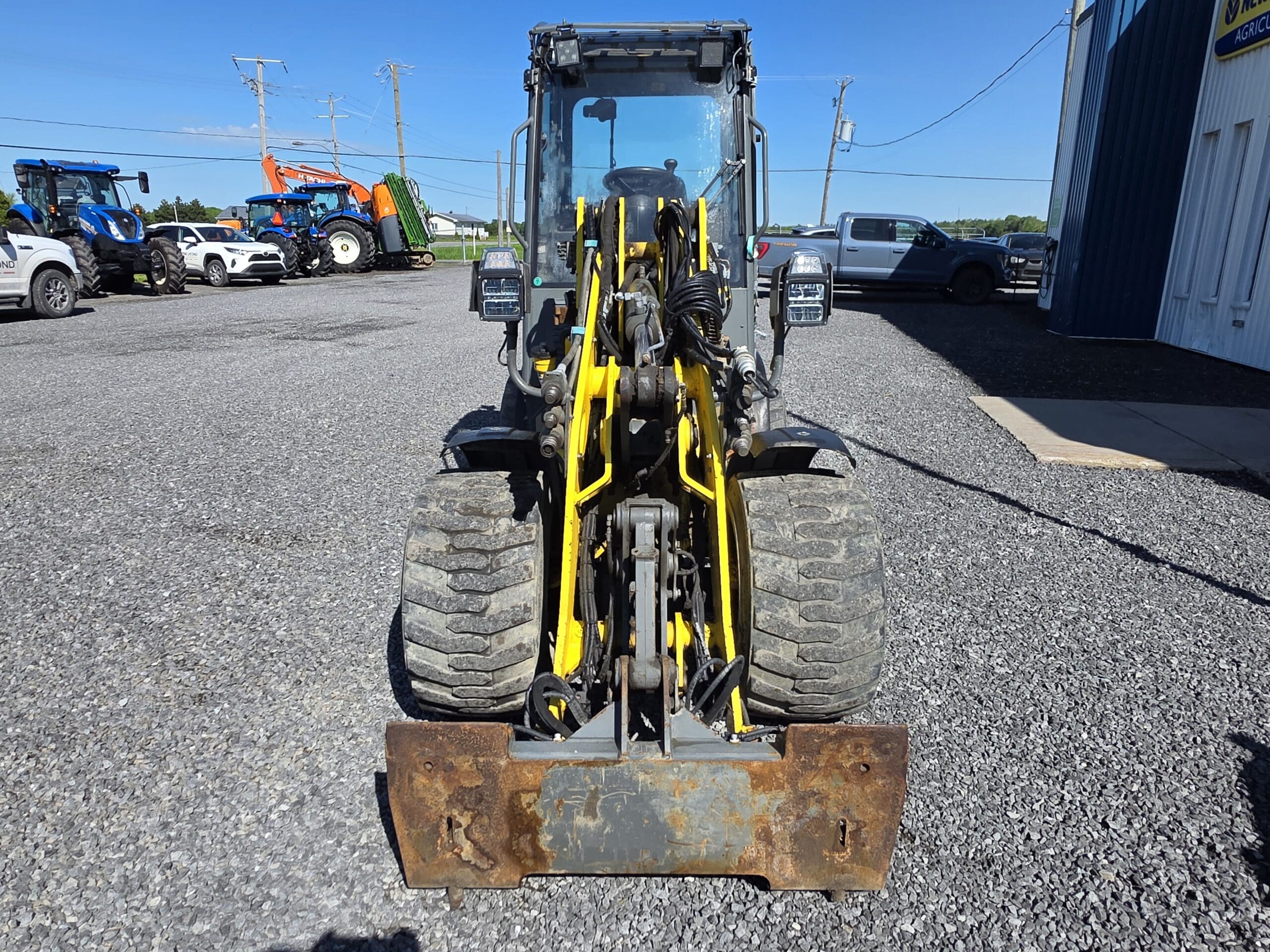 Vue de face d'une chargeuse sur pneus compacte avec des tuyaux hydrauliques jaunes, de gros pneus et un godet rouillé, garée sur du gravier à proximité d'autres véhicules et équipements agricoles sous un ciel clair.