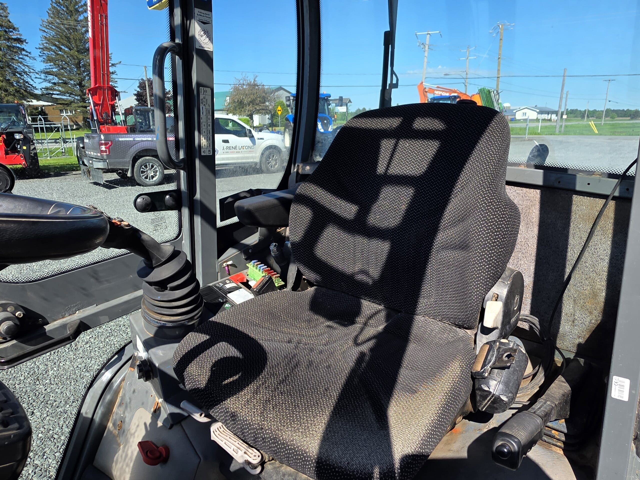 La photo montre le siège du conducteur et les leviers de commande à l'intérieur de la cabine d'un engin de chantier, avec des ombres projetées par le soleil sur le siège. L'équipement et les véhicules sont visibles à l'extérieur par la fenêtre de la cabine.