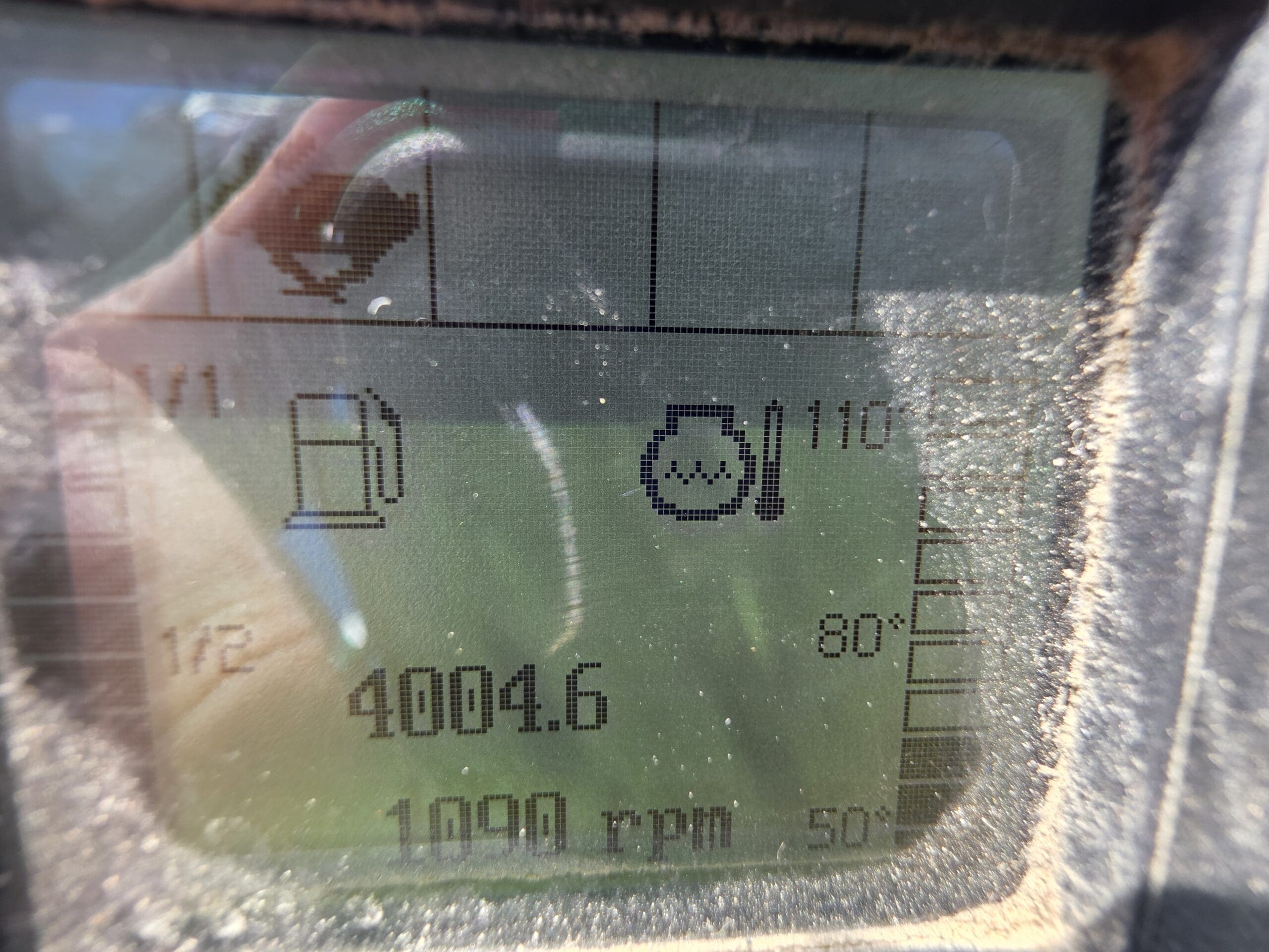 Un écran numérique poussiéreux affiche les jauges de carburant, de température du moteur, 4 004,6 heures et 1 090 tr/min. Des reflets et de la saleté obscurcissent partiellement l'écran.