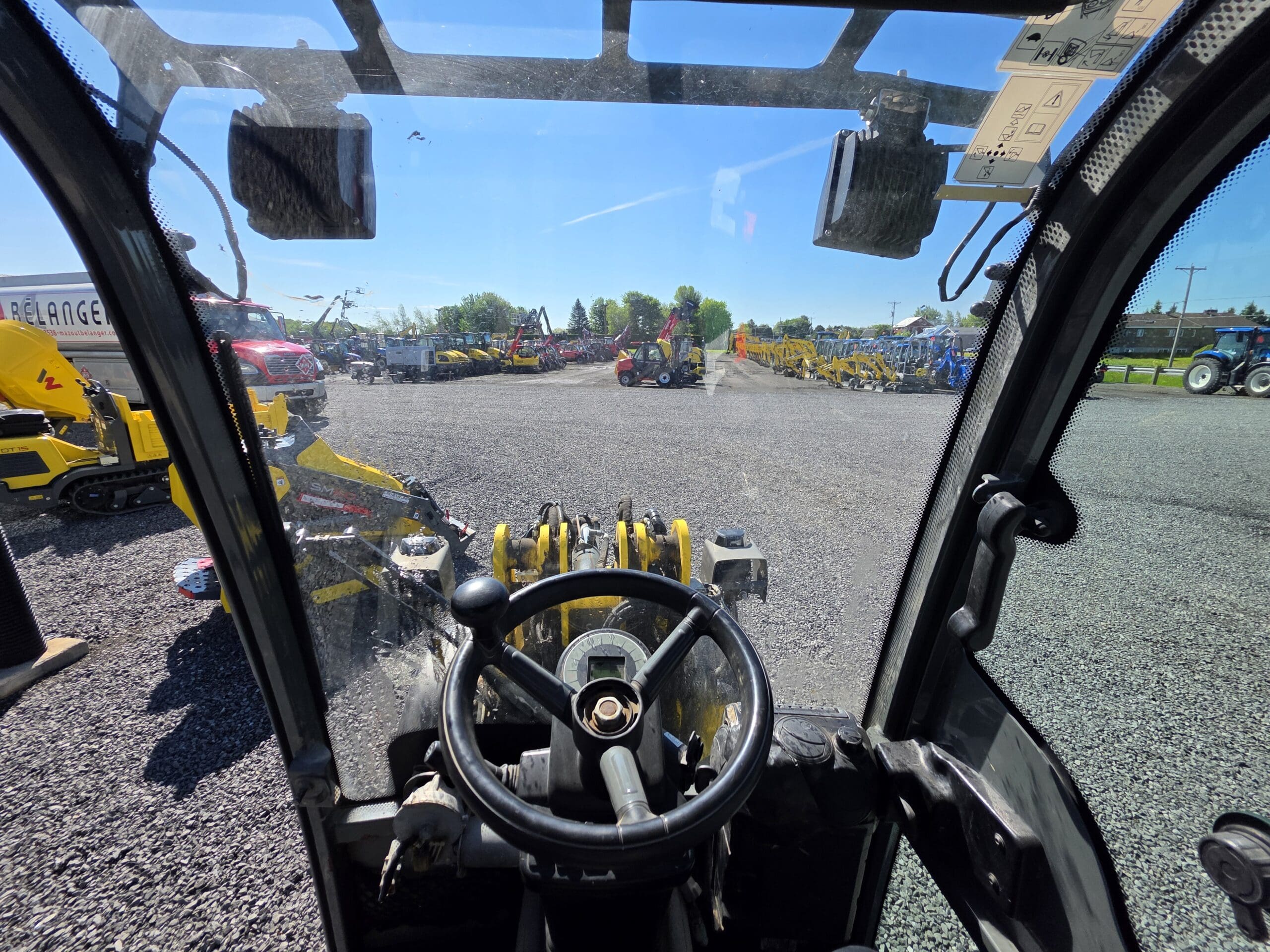 Vue depuis l'intérieur de la cabine d'un véhicule de construction, regardant à travers le pare-brise diverses machines jaunes et des personnes par une journée ensoleillée, avec du gravier et de l'équipement visibles à l'extérieur.