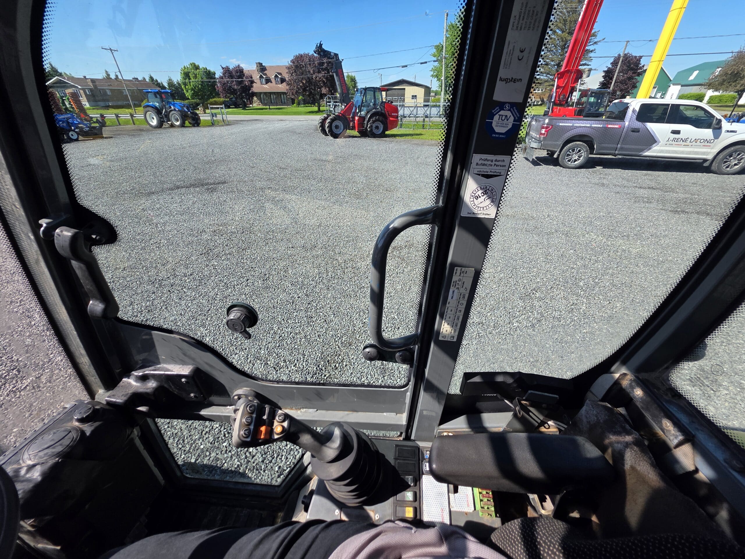 Vue de l'intérieur de la cabine d'un véhicule de construction, montrant le joystick de commande et l'accoudoir, regardant par la fenêtre des tracteurs, une camionnette et des bâtiments dans un terrain en gravier par une journée ensoleillée.
