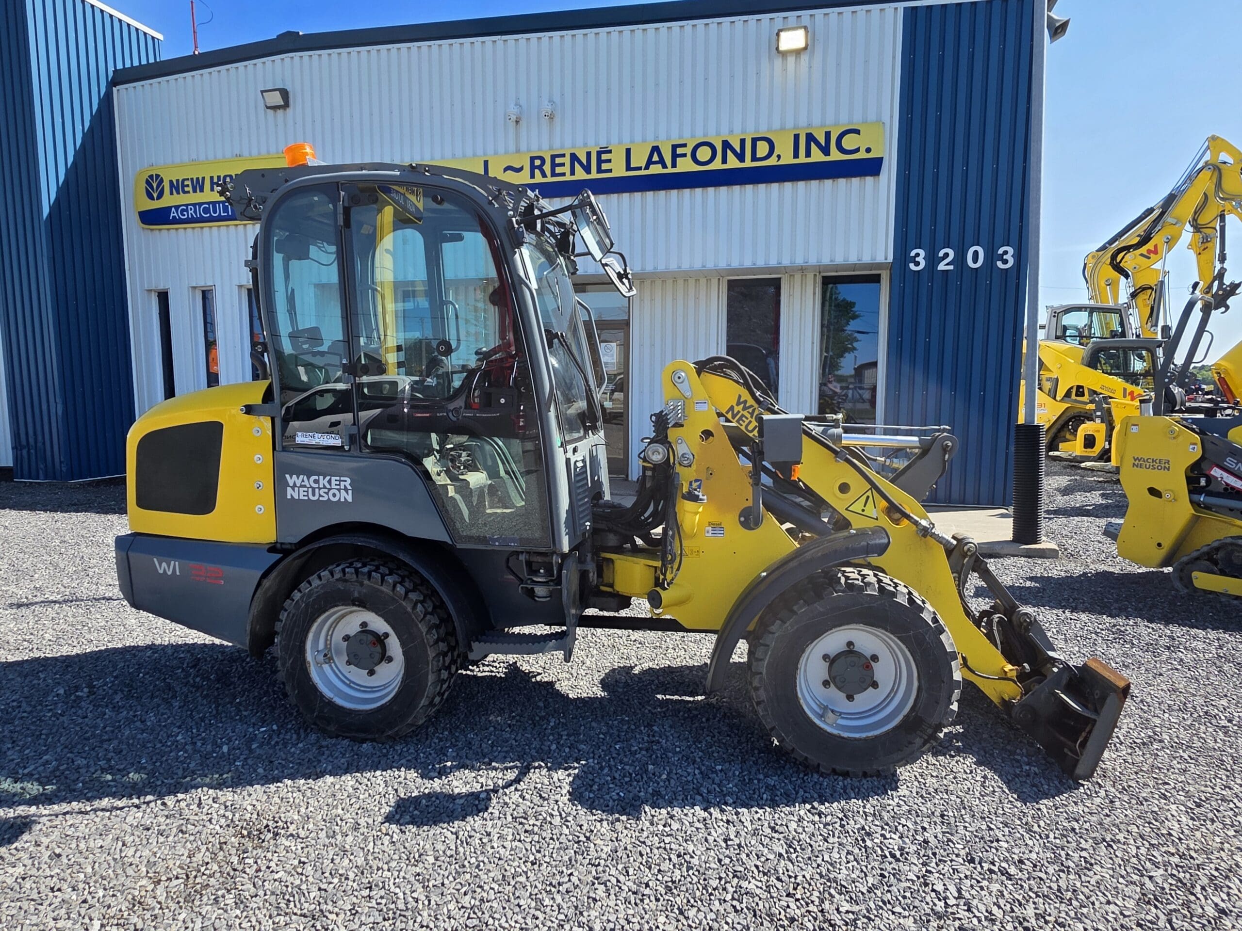 Une chargeuse sur pneus Wacker Neuson jaune et grise est garée sur du gravier devant un bâtiment bleu et blanc portant les inscriptions « René Lafond, Inc. » et « New Holland Agriculture ». D'autres engins de chantier sont visibles en arrière-plan.