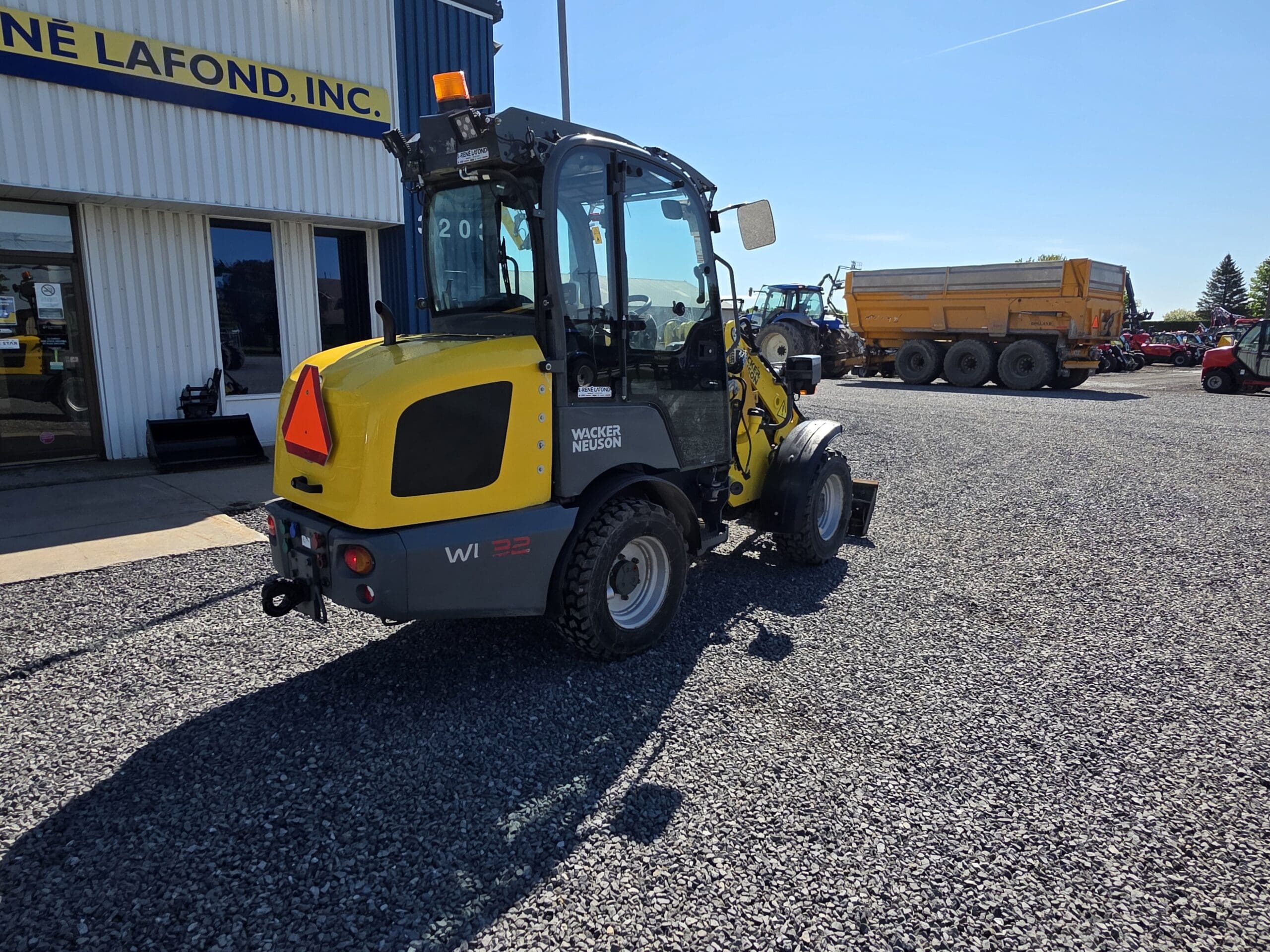 Une chargeuse sur pneus Wacker Neuson jaune est garée sur un terrain en gravier devant un bâtiment portant l'inscription « LAFOND, INC. ». Plusieurs gros véhicules sont visibles en arrière-plan par une journée ensoleillée.
