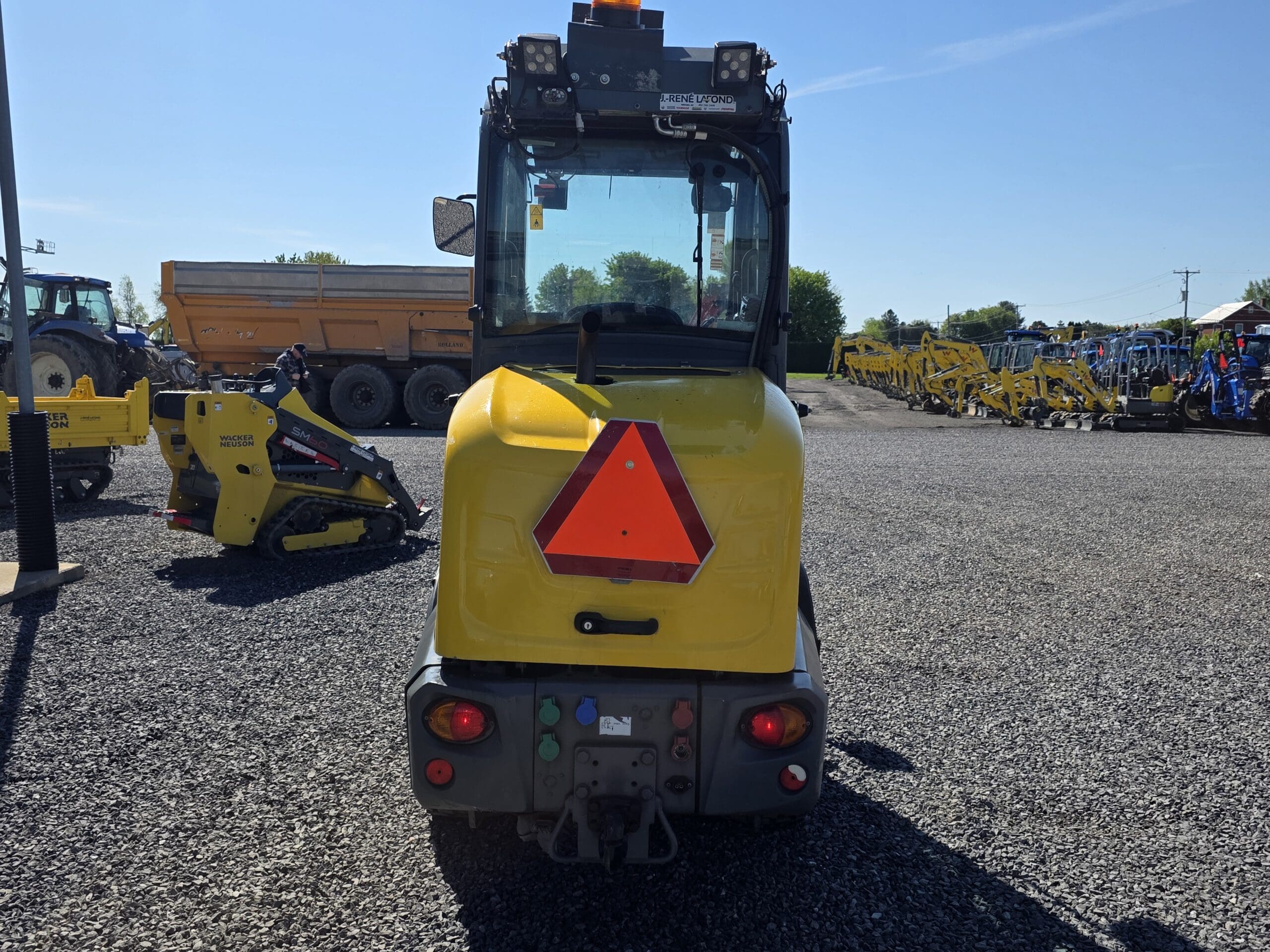 Un véhicule de construction jaune, vu de derrière, avec un triangle réfléchissant rouge, des boutons et des lumières ; d'autres machines et véhicules sont garés en arrière-plan sur un terrain en gravier sous un ciel ensoleillé.