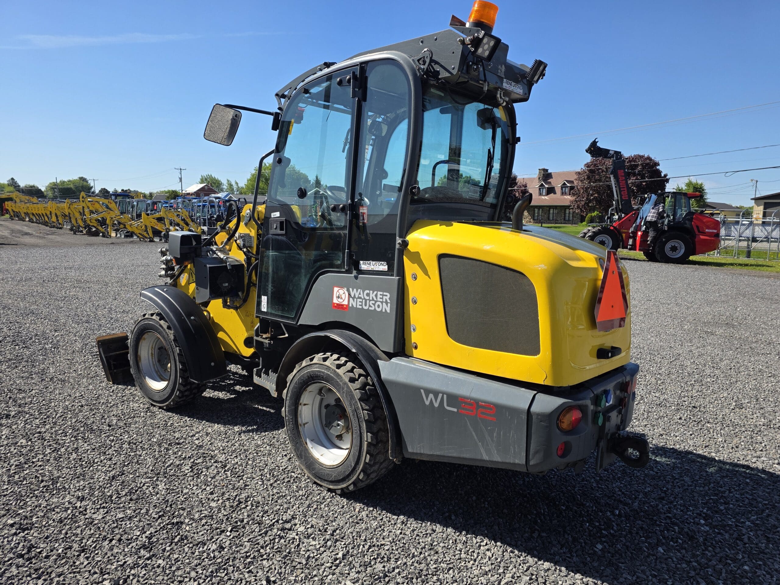 Une chargeuse sur pneus jaune Wacker Neuson WL32 est garée sur du gravier, avec des équipements de construction visibles en arrière-plan sous un ciel bleu clair.