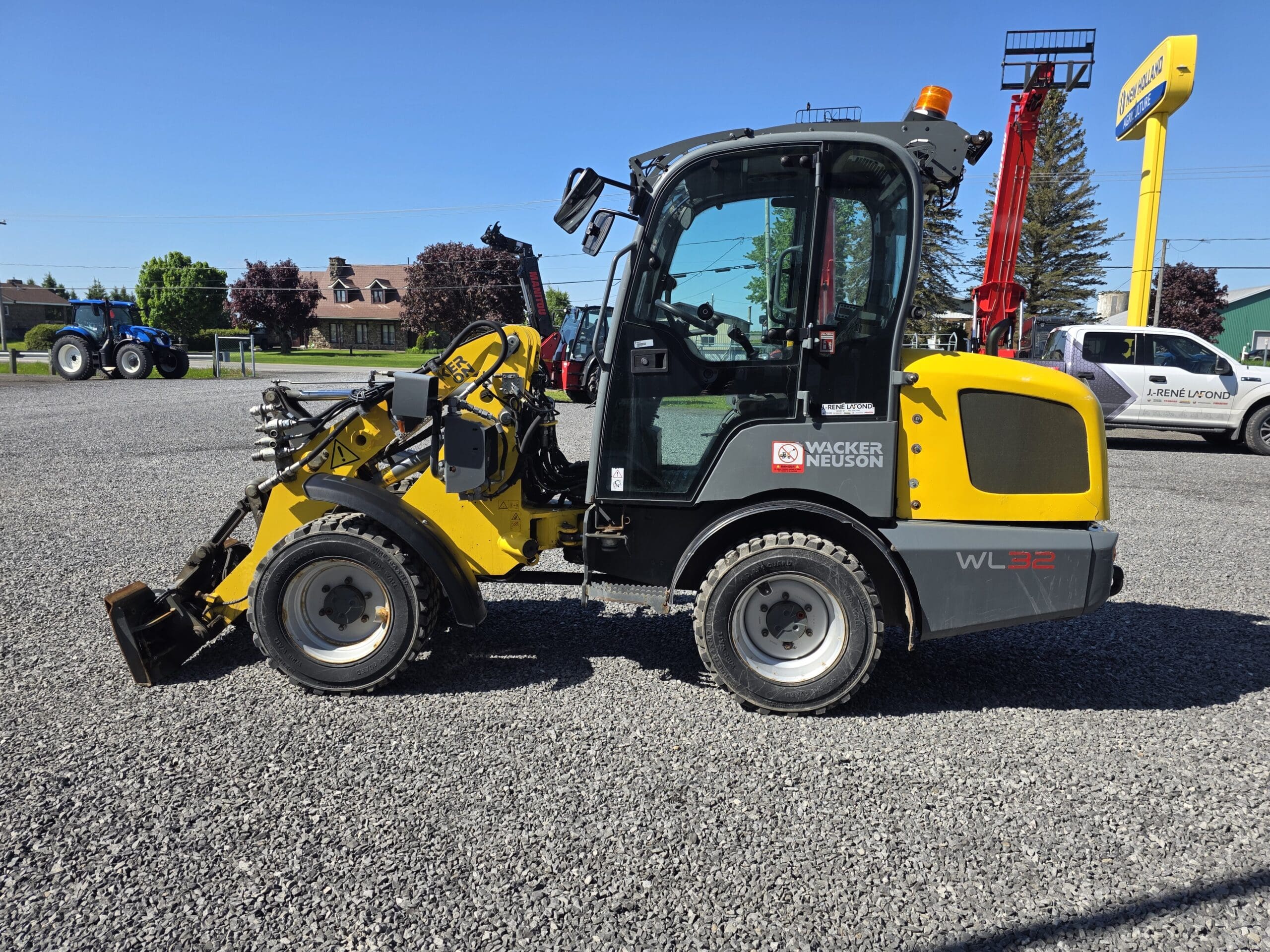 Une chargeuse compacte sur pneus Wacker Neuson WL32 jaune et grise garée sur un terrain en gravier, avec d'autres machines et arbres visibles en arrière-plan par une journée ensoleillée.